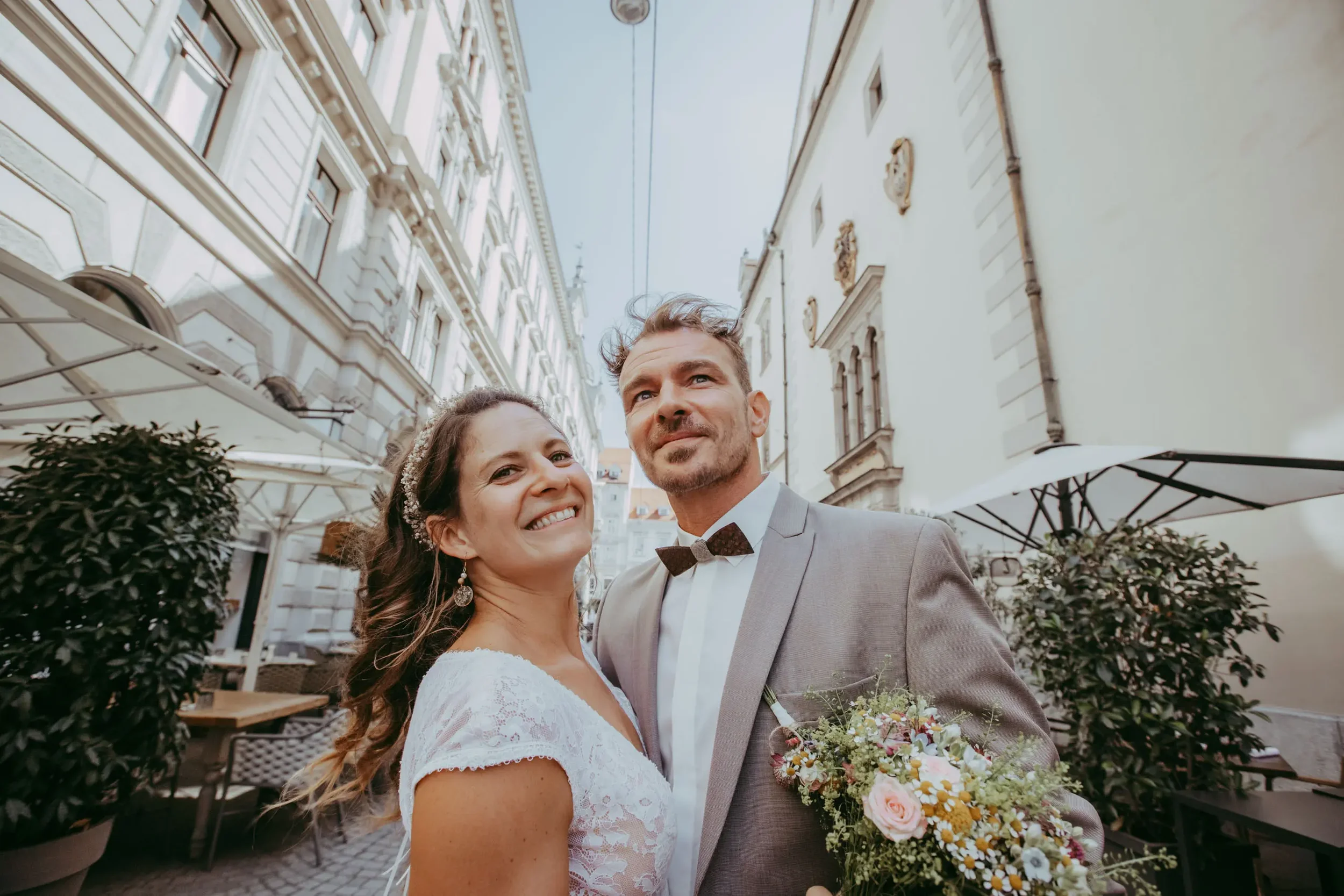 Ein Brautpaar macht ein Selfie in einer Stadtstraße. Die Frau trägt ein weißes Brautkleid mit Spitze und Ohrringe, der Mann trägt einen grauen Anzug mit Fliege. Sie halten einen Brautstrauß mit Blumen in Pastellfarben. Im Hintergrund sind alte Gebäud