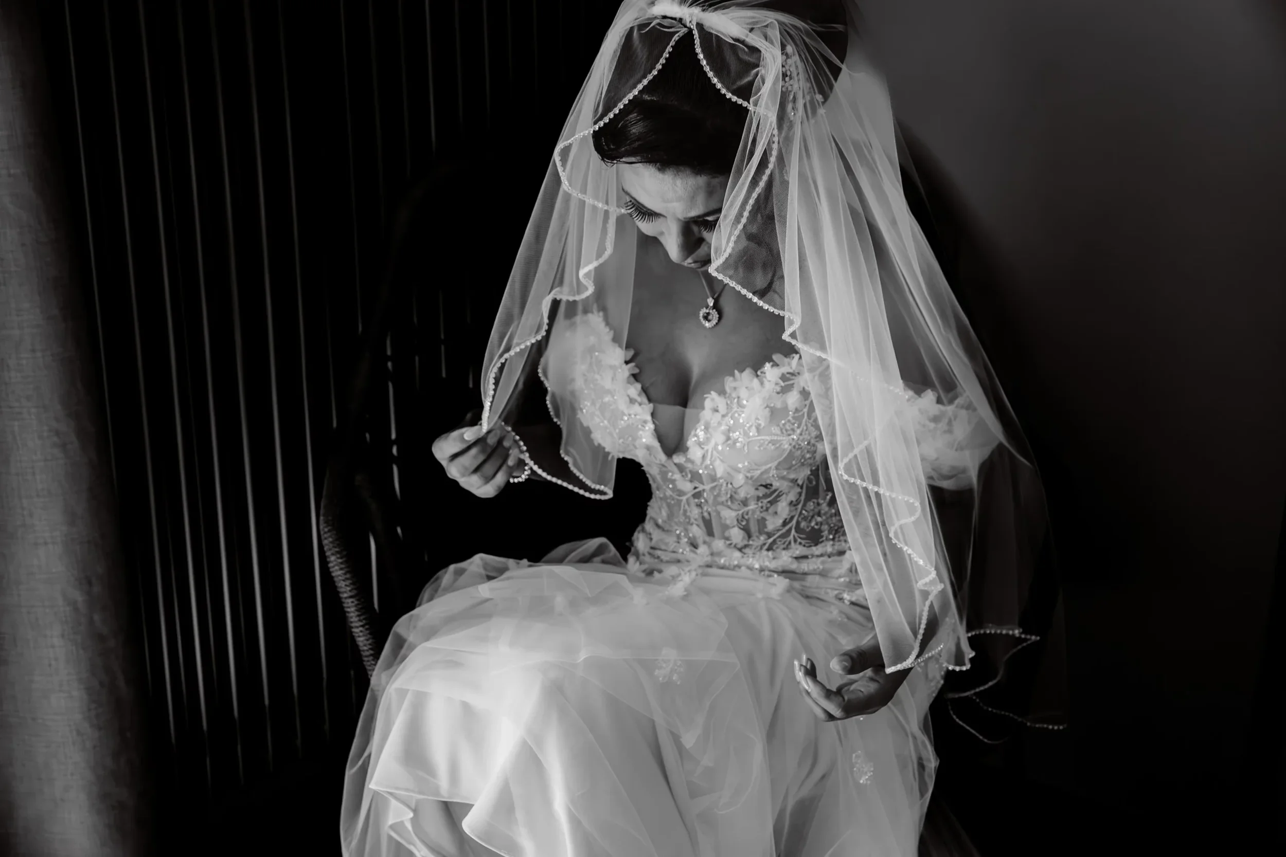 Schwarz-Weiß-Foto einer Braut in Hochzeitskleid mit Schleier, die einen Schmuck hält und nach unten blickt.