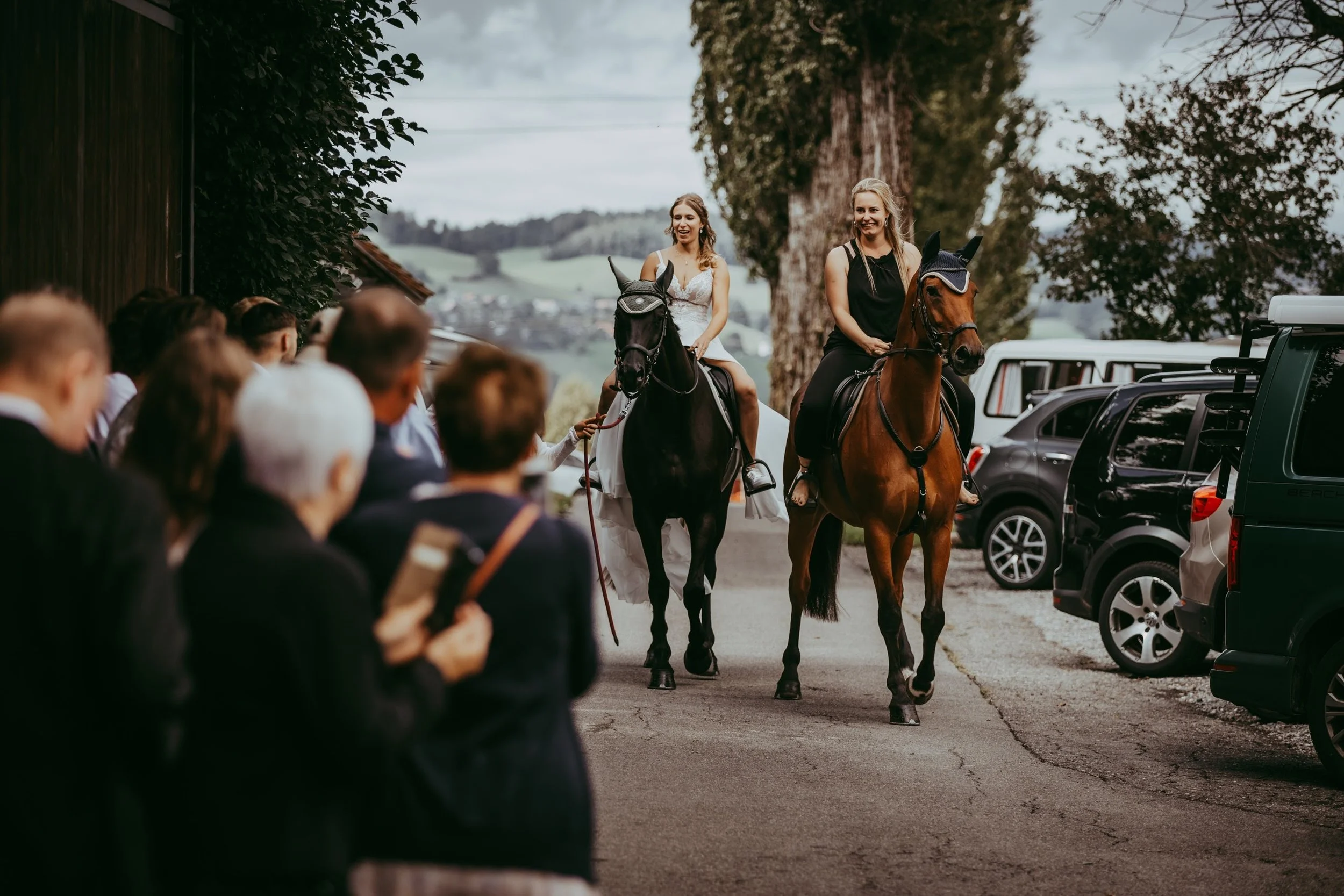 Zwei Frauen reiten auf Pferden vor einer Menschenmenge bei einer Veranstaltung im Freien, umgeben von parkenden Autos, Bäumen und Hügeln im Hintergrund.