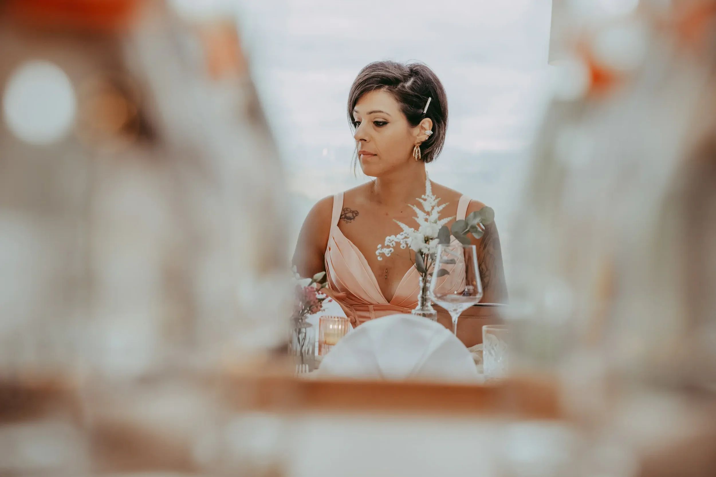 Eine Frau mit dunklem, kinnlangem Haar, die elegant gekleidet ist und auf einem festlich gedeckten Tisch sitzt, umgeben von Blumen und Kerzen.