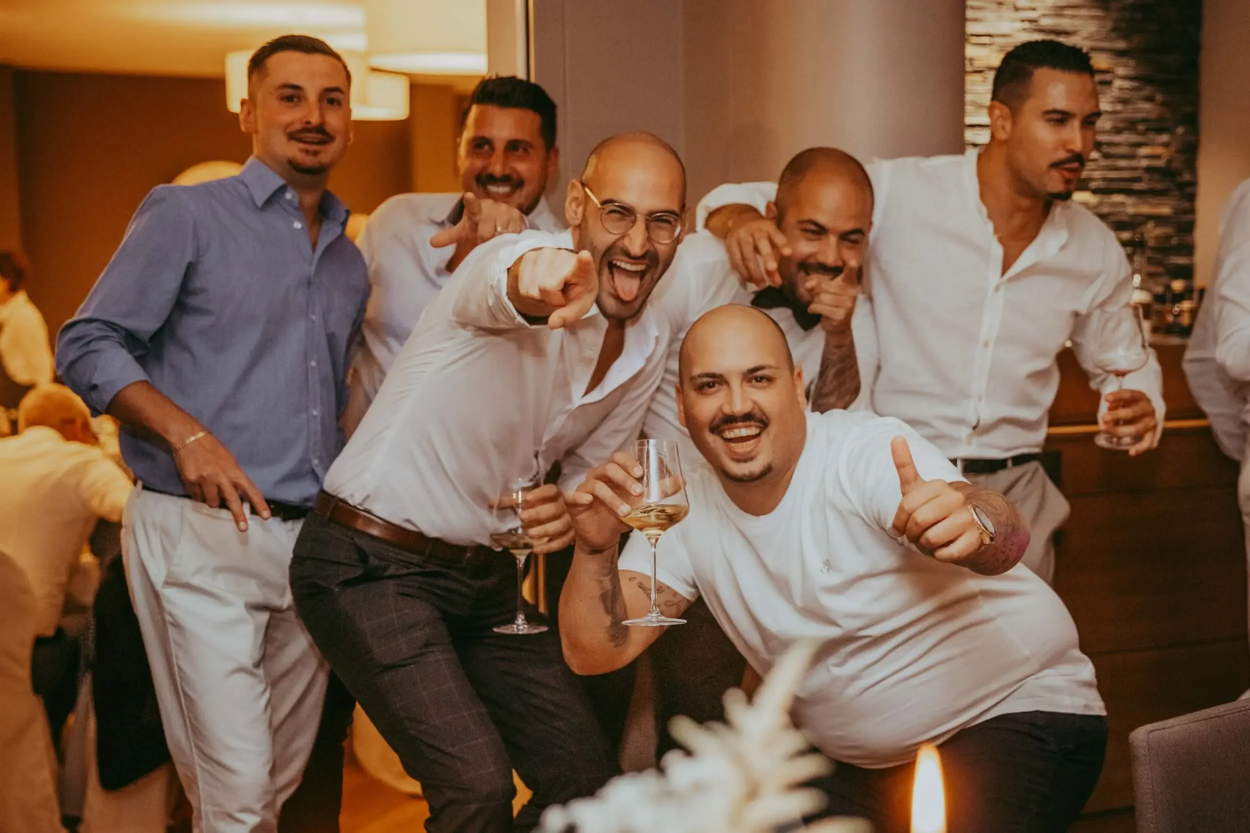 Eine Gruppe von sechs Männern, die bei einer Feier lachen, anstoßen und Spaß haben, einige halten Weingläser.