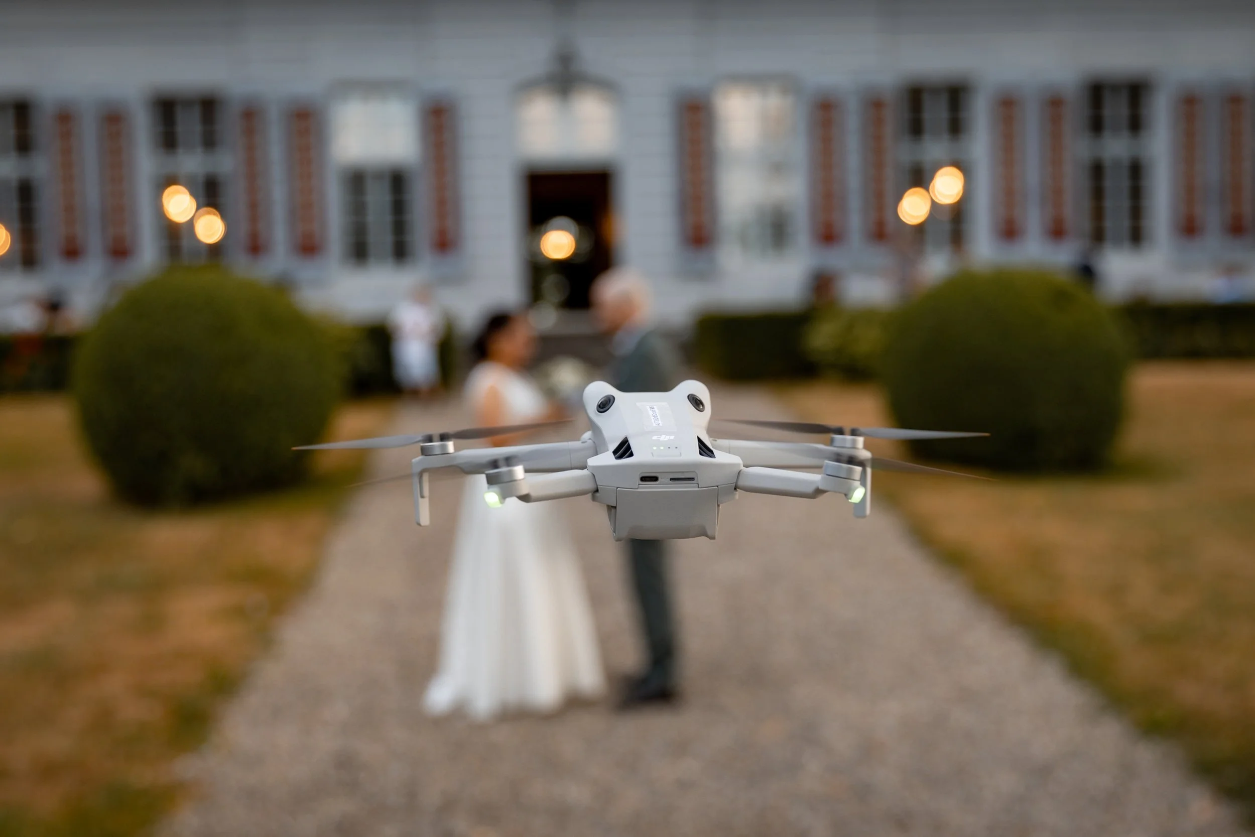 Eine Drohne fliegt auf einem Weg vor einem Hochzeithaus, im Hintergrund sind zwei Menschen in Hochzeitskleidung zu sehen.