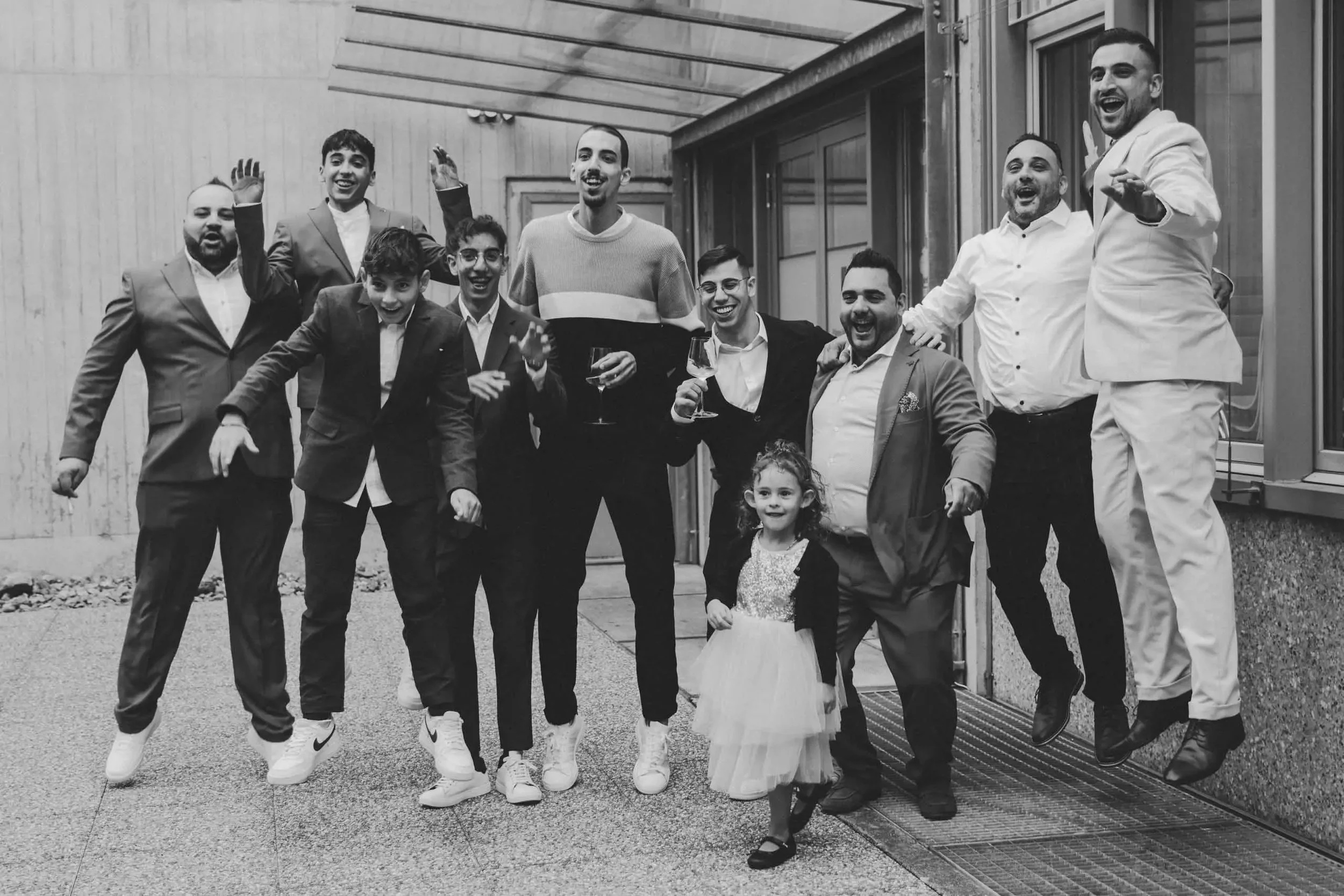Gruppe von fröhlichen Menschen, die auf einer Terrasse stehen, einige heben die Hände und halten Weingläser. Es sind Männer, Frauen und Kinder, alles in Festtagskleidung.