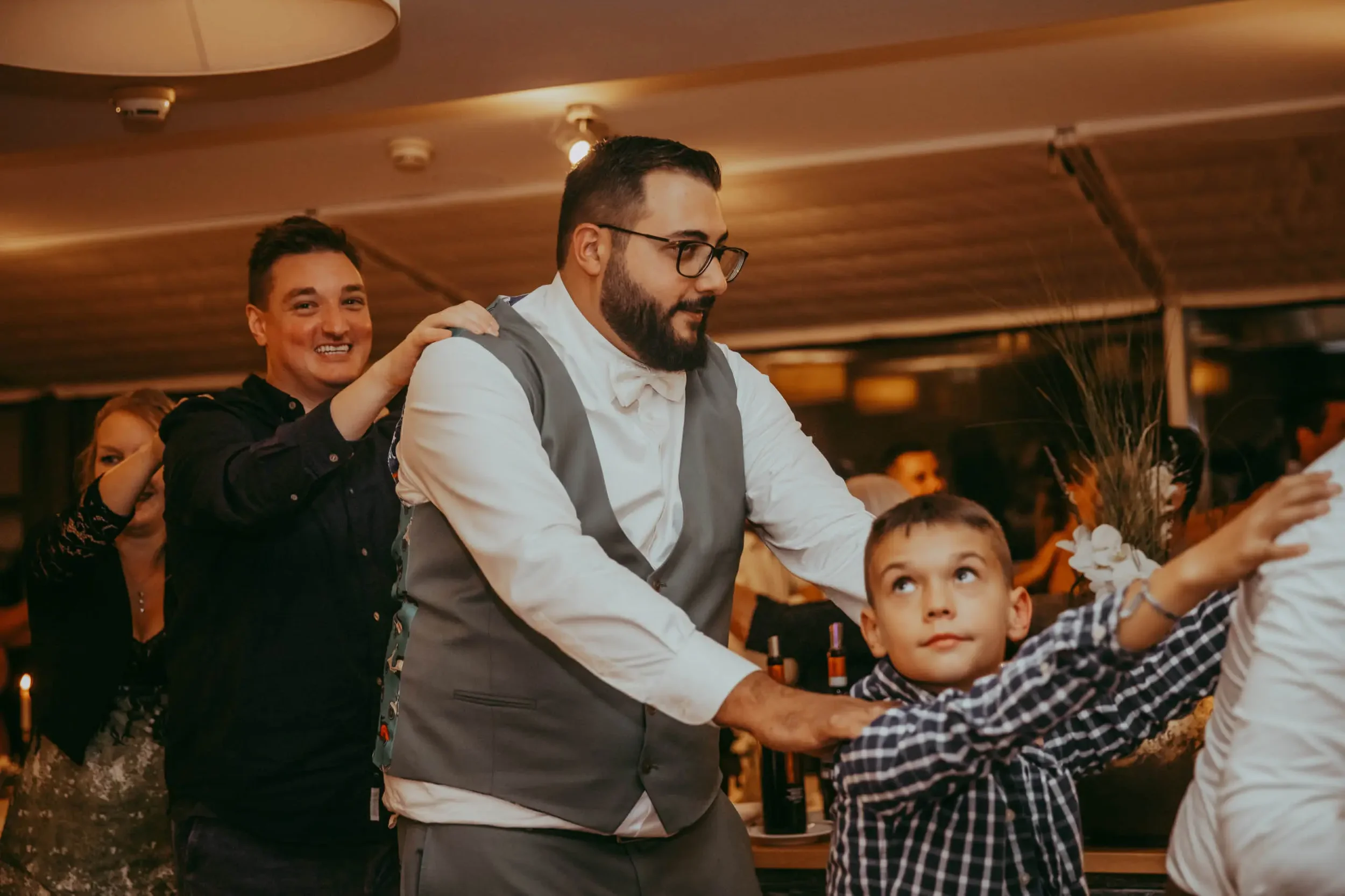 Menschen bei einer Feier, eine Person mit Brille und Bart in einem Anzug, Umschlungen mit einem jungen Jungen, im Hintergrund weitere Gäste in einem festlich dekorierten Raum.