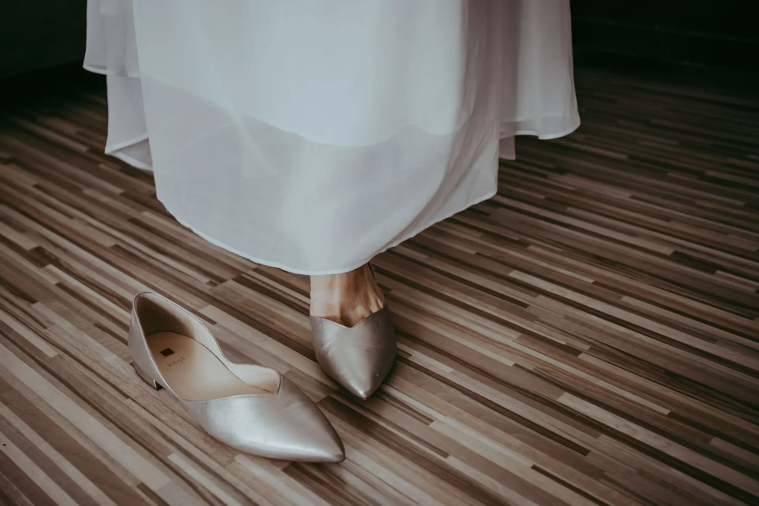 Ein Paar goldene High Heels stehen auf einem Holzparkett, daneben eine Frau in einem weißen Kleid. Ein weiteres Paar goldene Schuhe liegt neben den Heels.