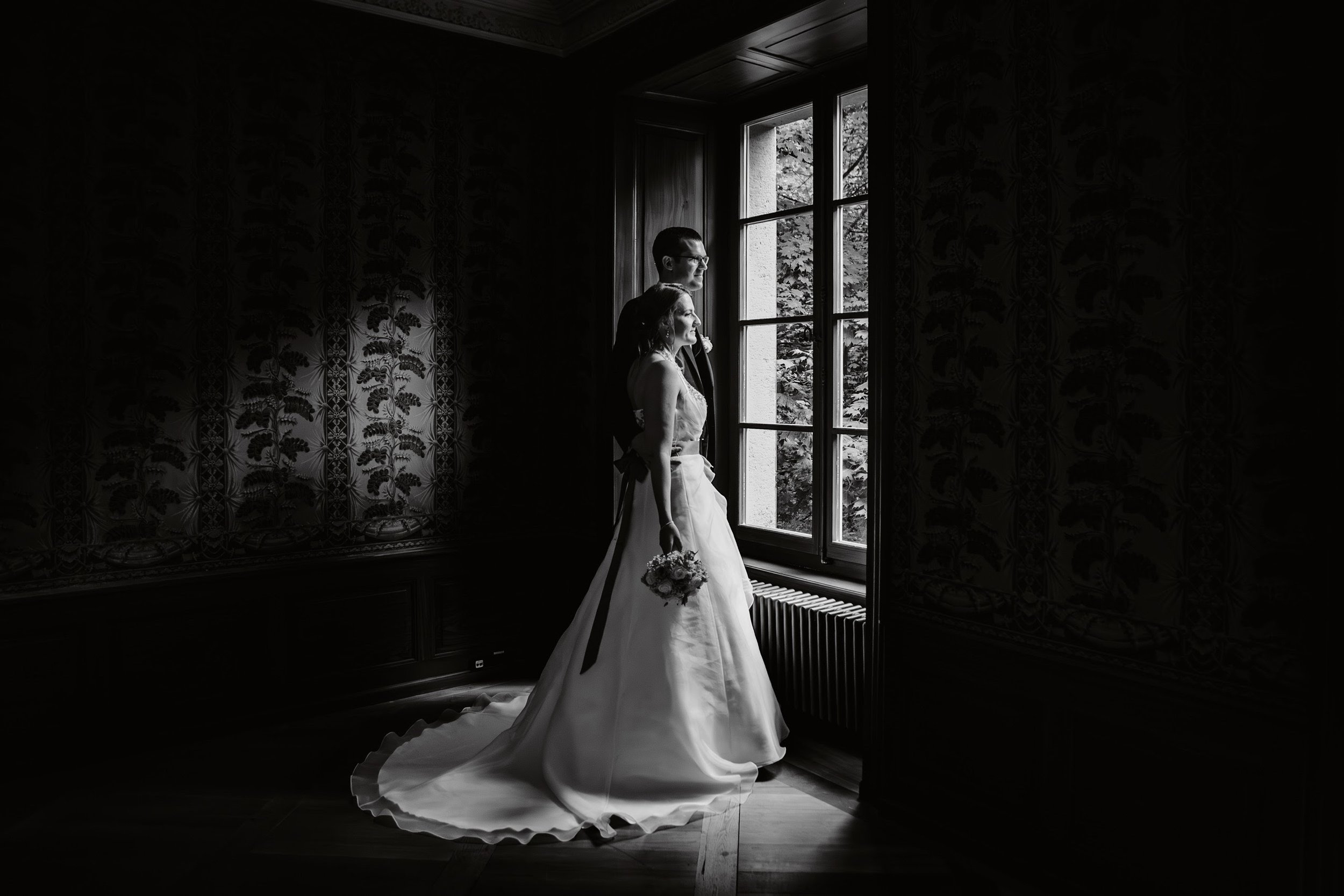 Braut und Bräutigam stehen im Raum an einem großen Fenster, im Schwarz-Weiß-Foto. Die Braut trägt ein langes, schulterfreies Hochzeitskleid und hält einen Blumenstrauß, der Bräutigam trägt einen dunklen Anzug und eine Brille. Draußen sind Bäume zu se