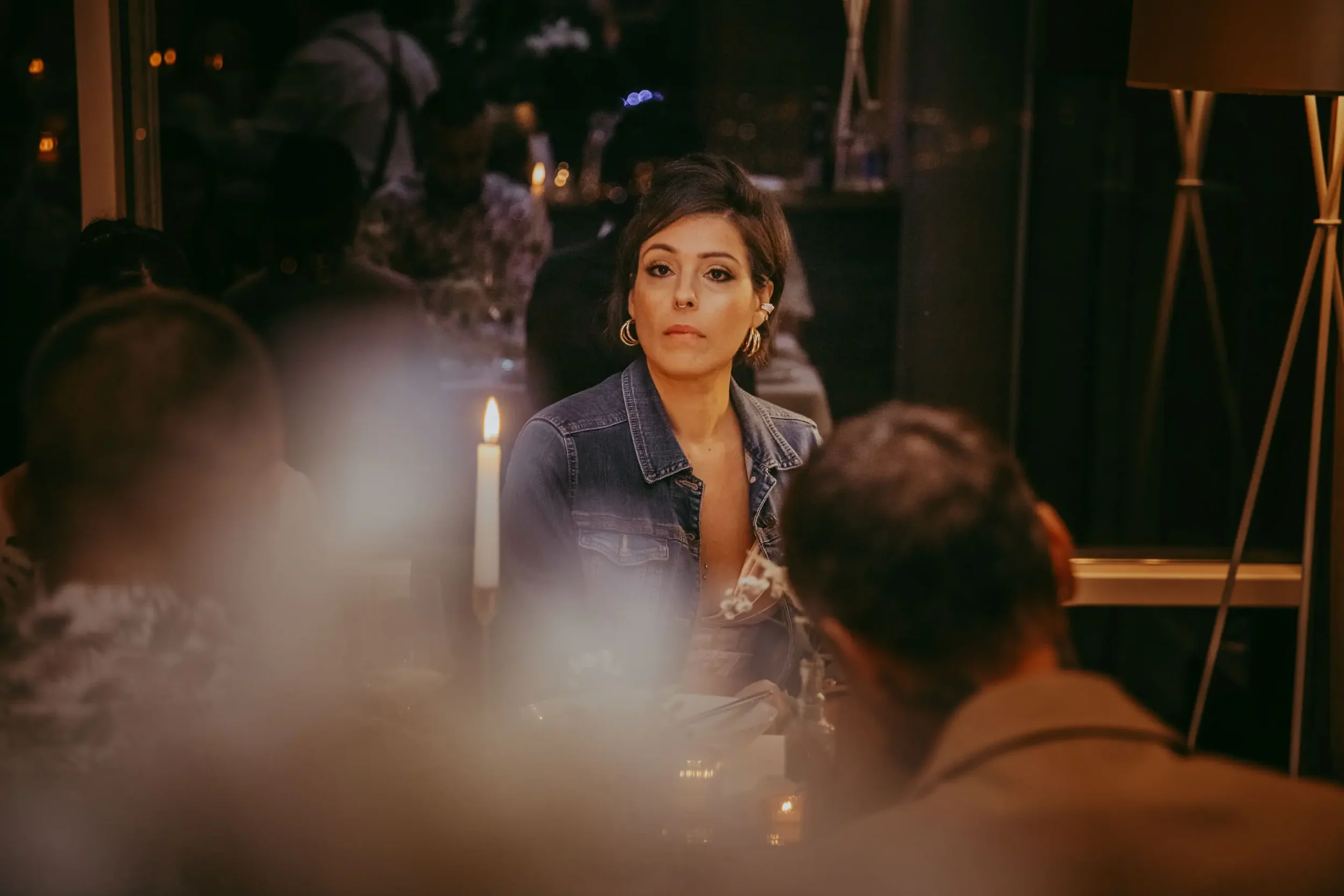 Eine Frau mit dunklen Haaren, Ohrschmuck und einem Jeansjacke sitzt an einem Tisch in einem Restaurant bei Kerzenschein.