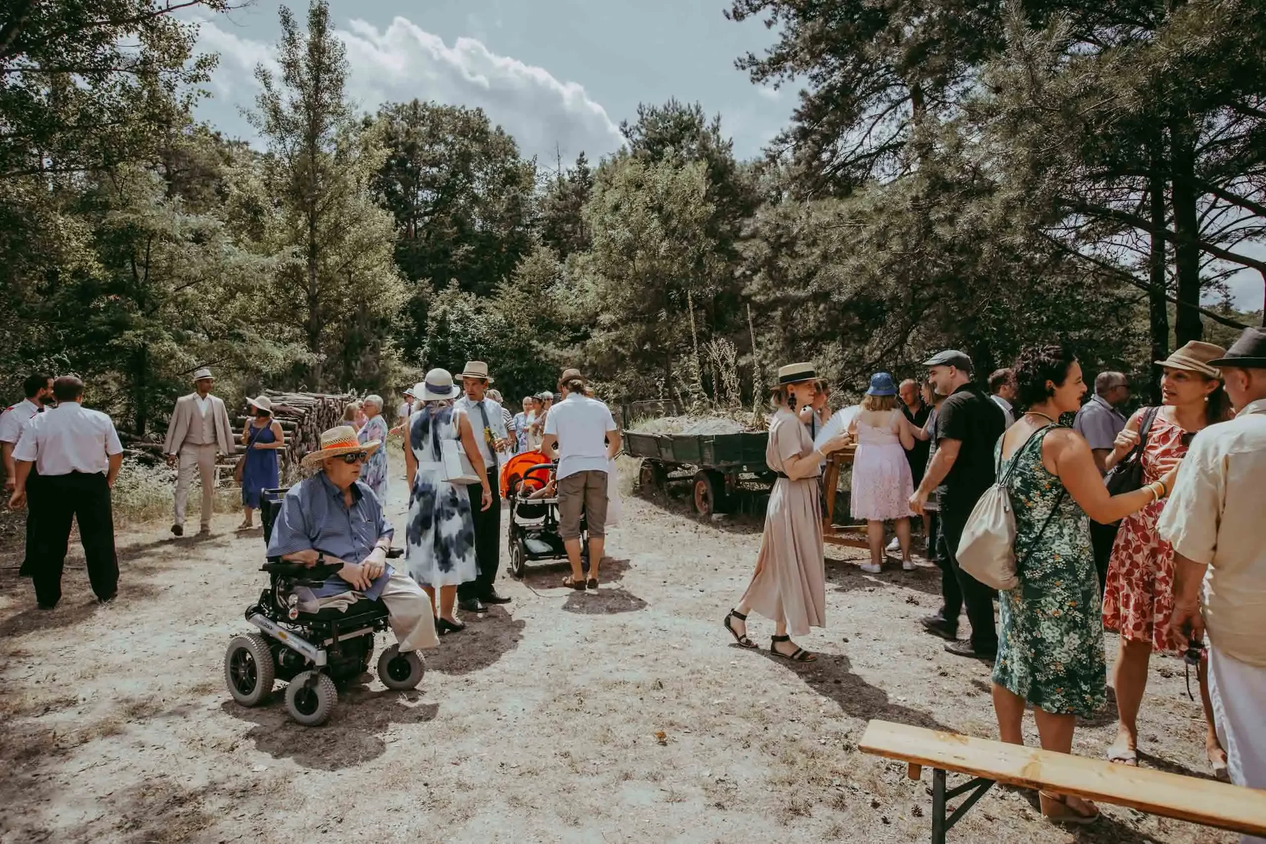 Eine Gruppe von Menschen, darunter ältere Personen, junge Erwachsene und eine Person im Rollstuhl, trifft sich im Freien in einem bewaldeten Gebiet an einem sonnigen Tag. Viele tragen Hüte und sommerliche Kleidung.