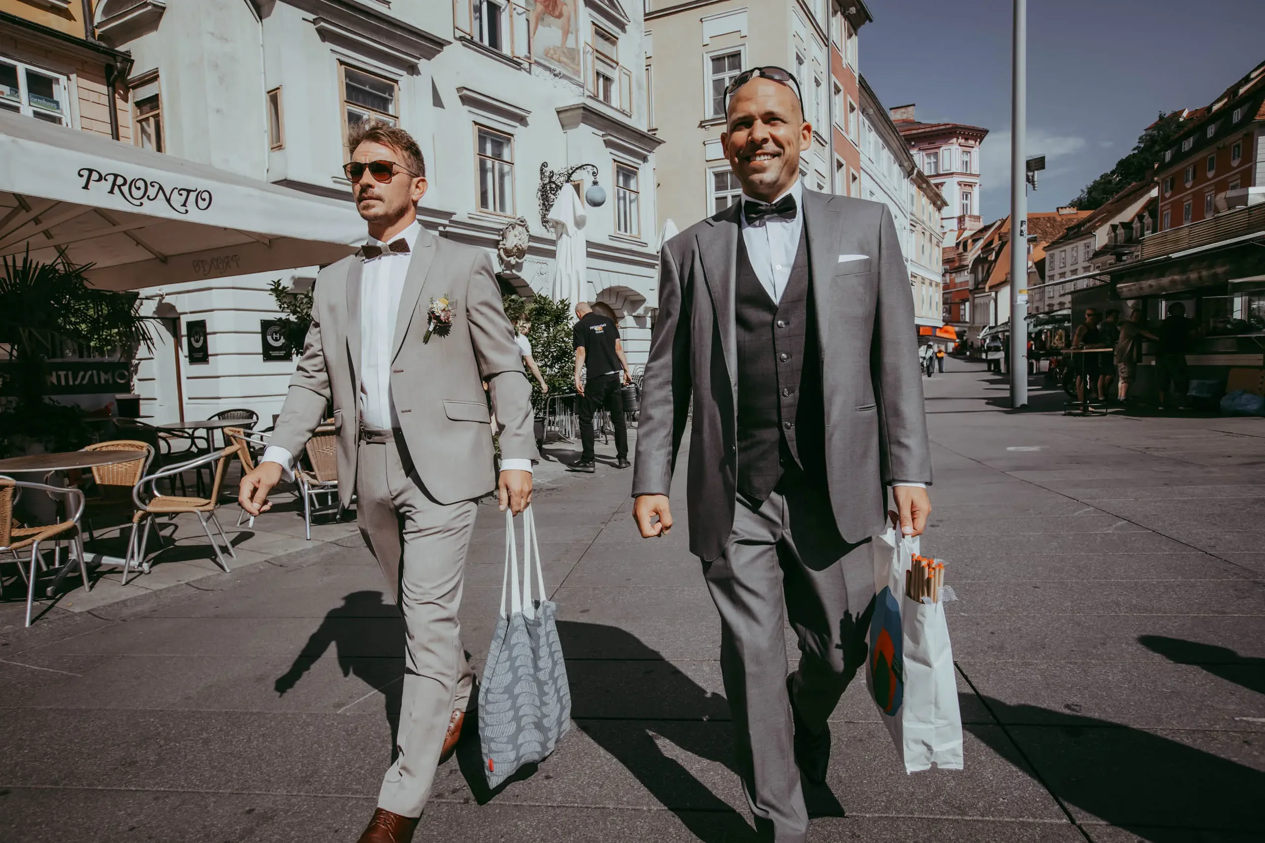 Zwei Männer in Anzügen und Krawatten gehen auf einer Straße in einer Stadt, rechts trägt einer Sonnenbrille. Im Hintergrund sind alte Gebäude, eine Gastronomie mit ein paar Tischen im Freien und einige Leute sichtbar.