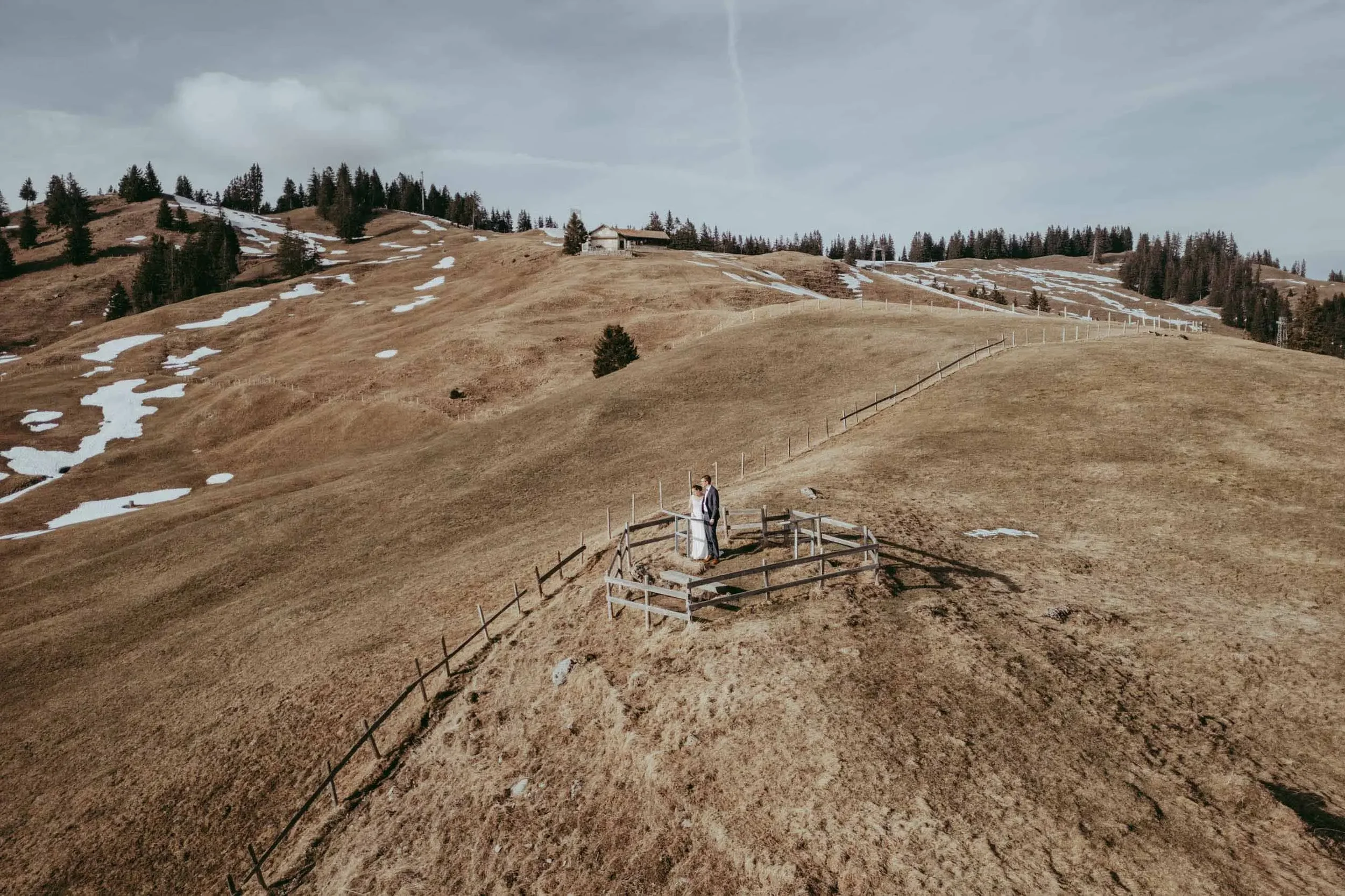 Ein Brautpaar steht auf einer Holzplattform in einer bergigen, kahlen Landschaft mit wenigen Bäumen, bei bewölktem Himmel.