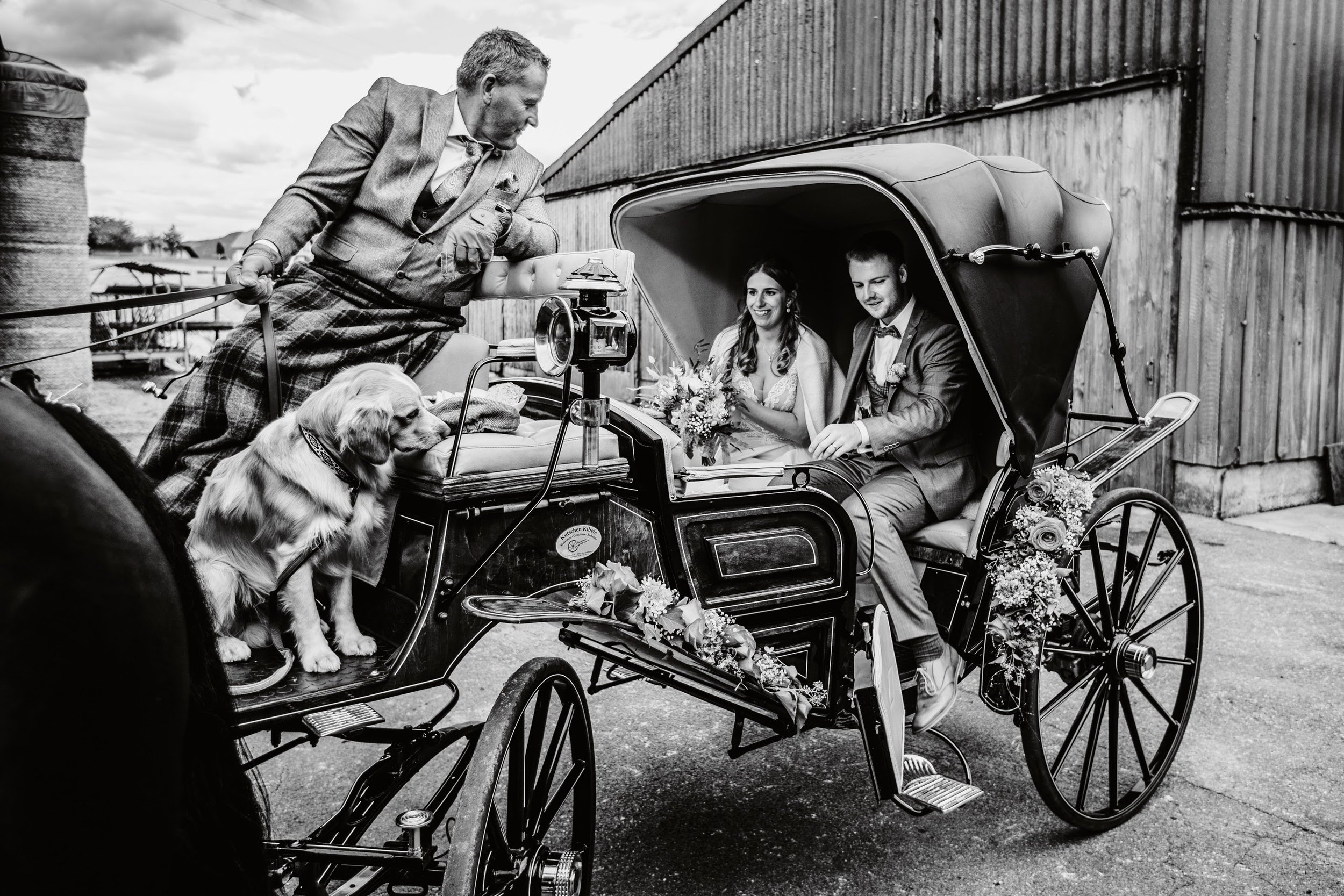 Brautpaar in Hochzeitsszene in einem vordernden kleinen roten Kutschwagen, der mit Blumen geschmückt ist, während ein Mann in traditioneller Kleidung den Wagen wegzieht, mit Hund auf der Kutsche und eine Hochzeitsfrau, die im Hintergrund lächelt.