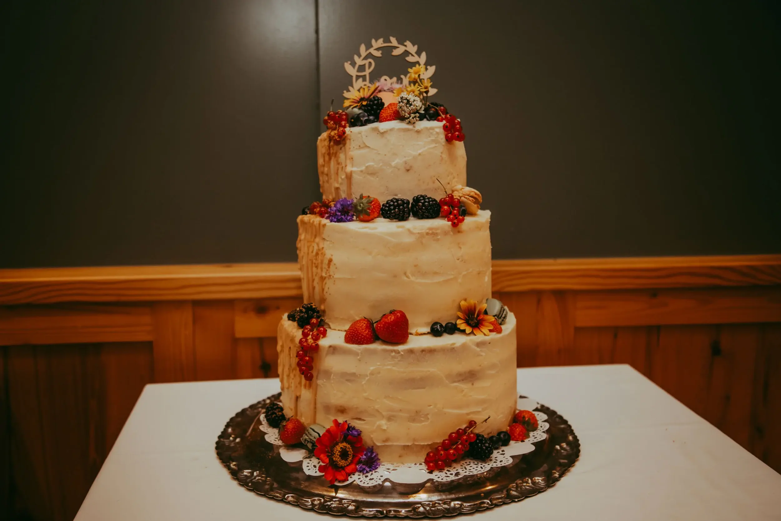 Mehrstöckige Hochzeitstorte mit Creme-Fassade, dekoriert mit frischen Beeren, Blumen und essbarem Dekor, auf einer silbernen Tortenplatte.