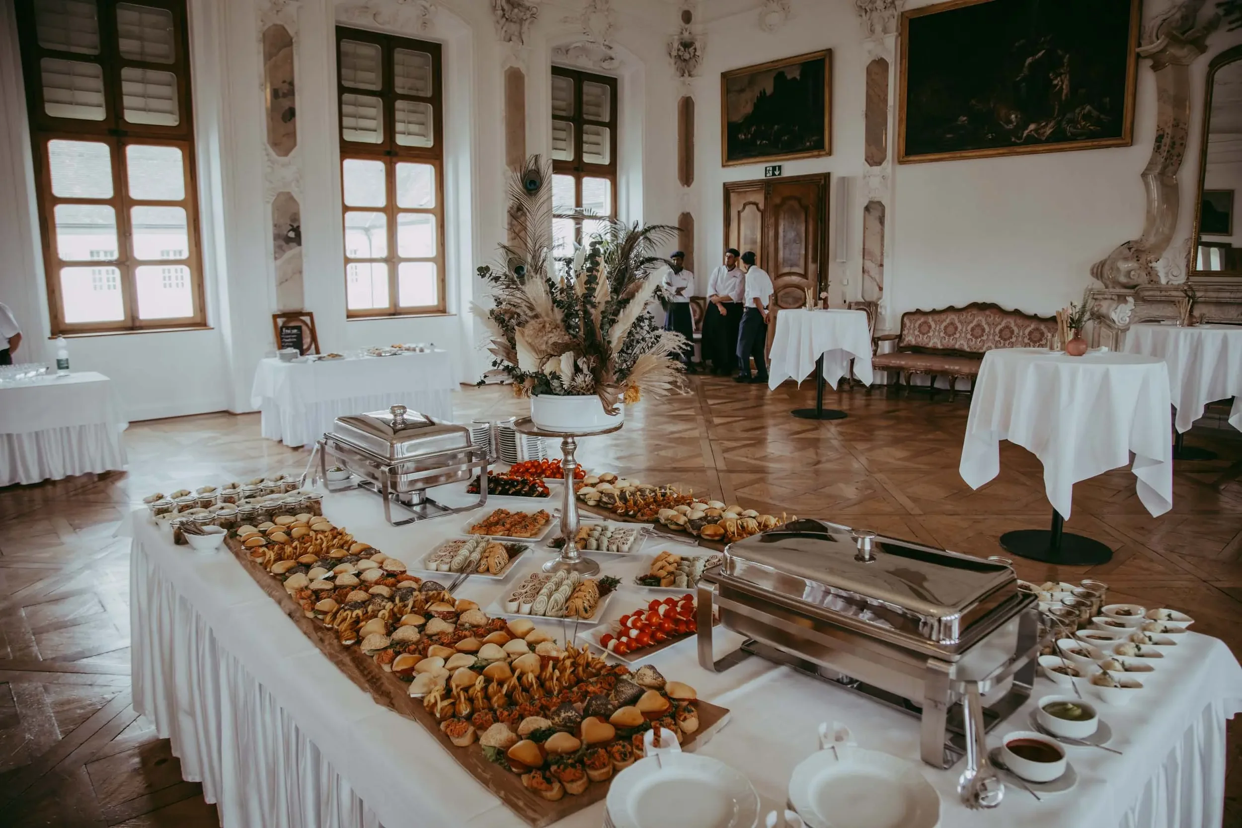 Ein festlich gedeckter Buffet-Tisch im eleganten Saal mit großen Fenstern, Blumen und Gebäck, flankiert von Kellnern.