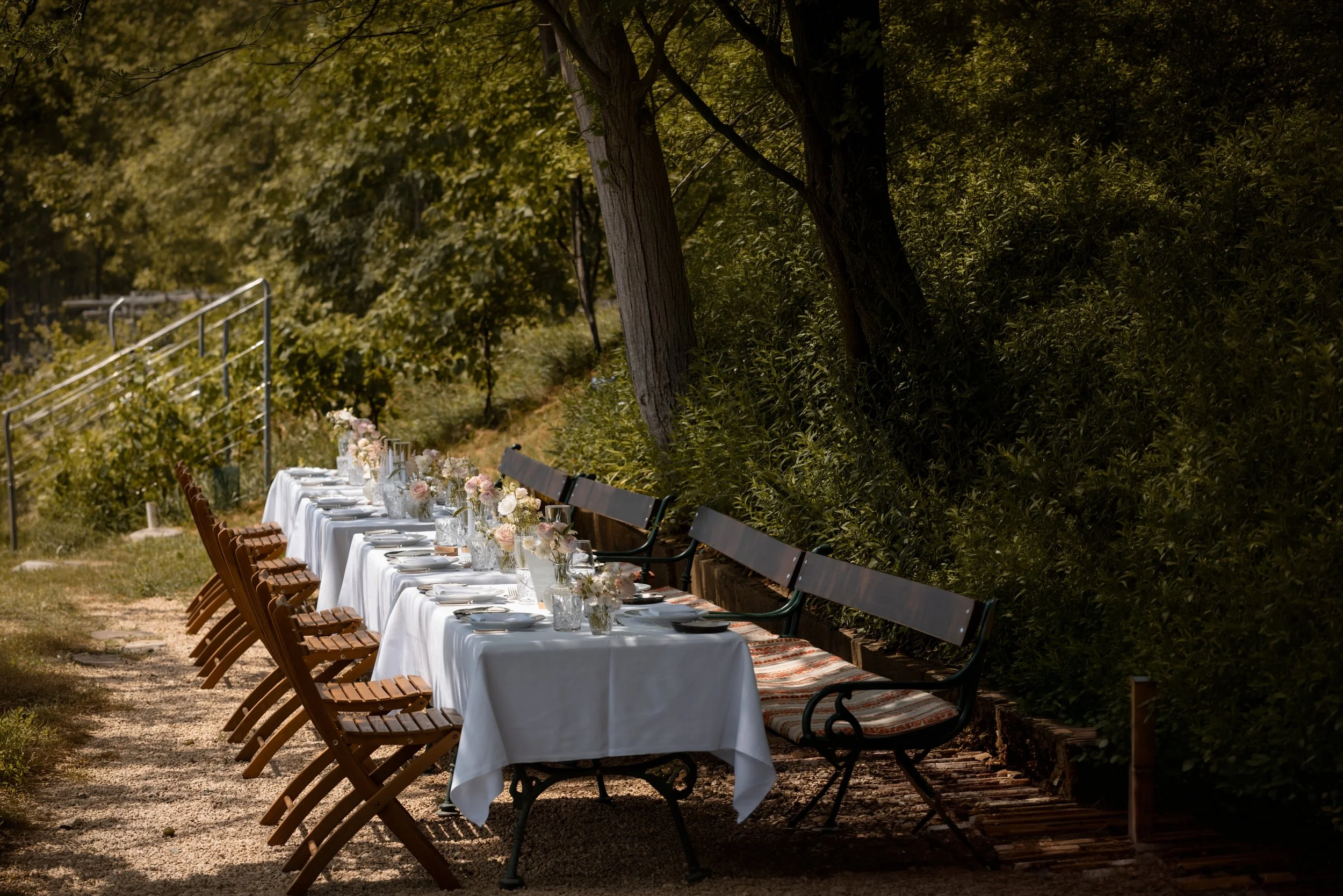Ein länglicher Gartentisch mit weißen Servietten und Blumenarrangements, umgeben von Holz- und Metallbänken, im Schatten von Bäumen, für eine outdoor Feier vorbereitet.