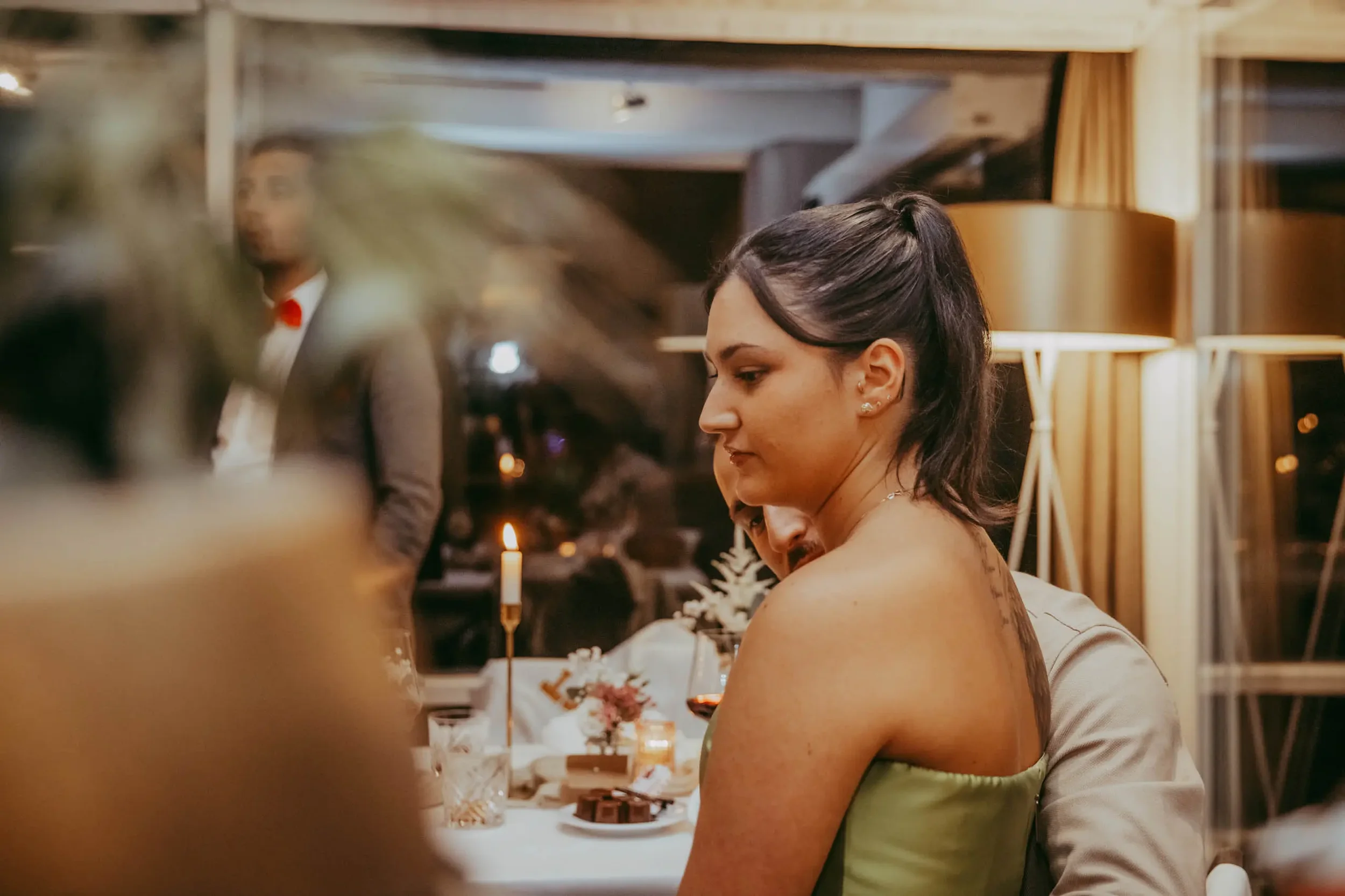Junge Frau mit dunklen Haaren und Piercings, die bei einer Feier mit Kerzen und Blumen sitzt, im Hintergrund sieht man einen Mann im Anzug.