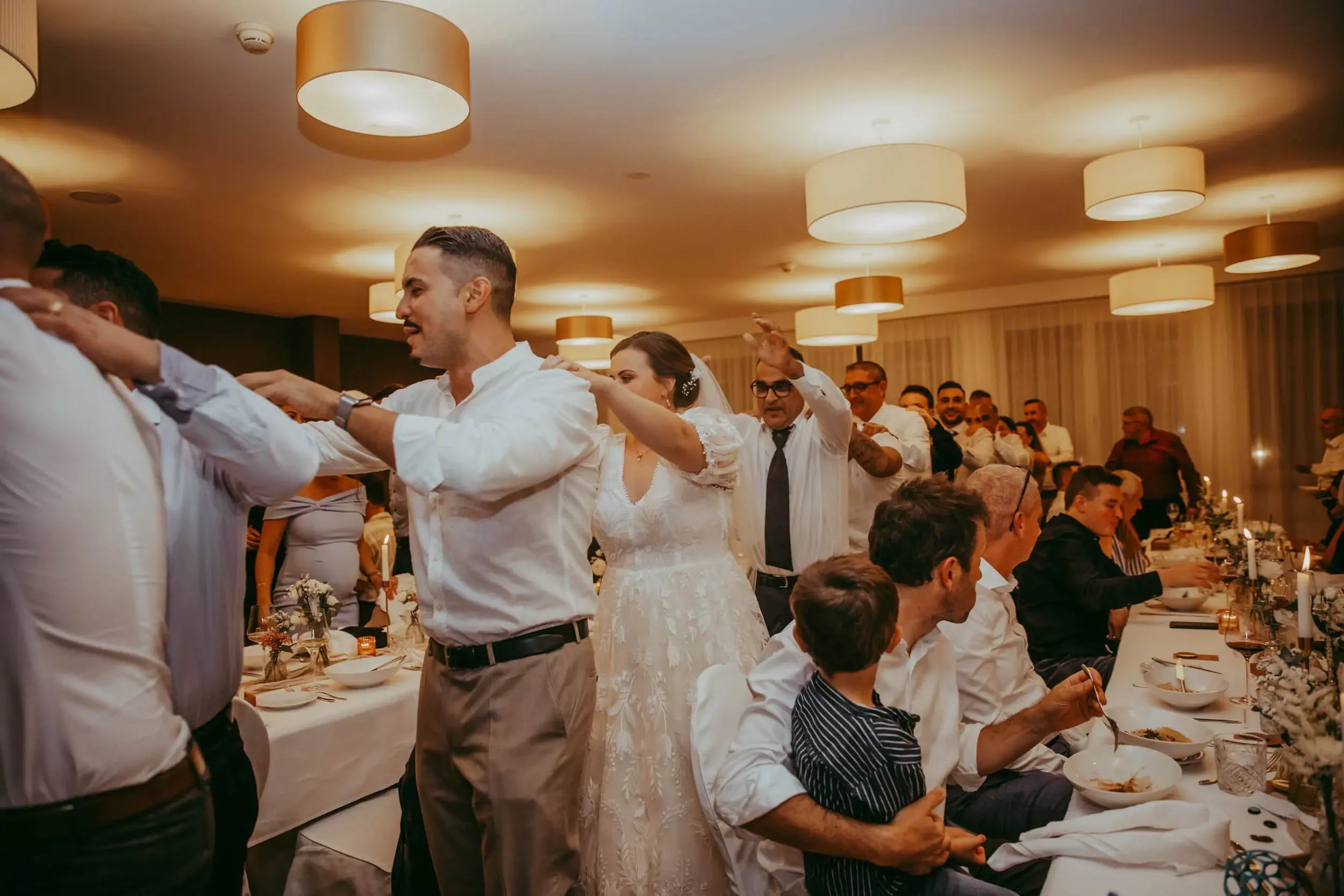 Hochzeitsszene mit Tanzpaaren und Gästen in einem festlich gedeckten Raum mit runden Lampen an der Decke.