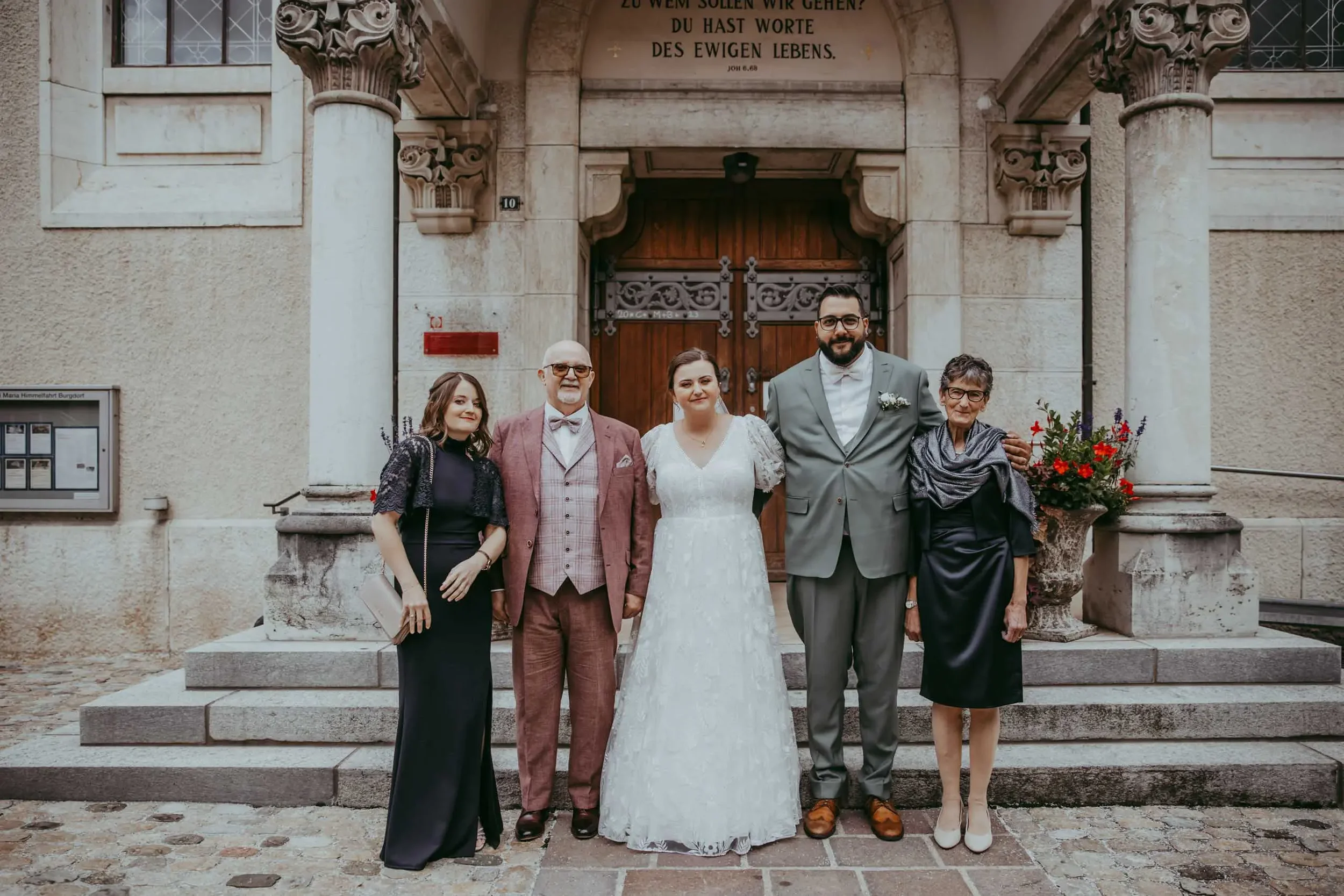 Eine Gruppe von sechs Menschen, darunter ein Paar in Hochzeitskleidung, steht vor einer alten Steinarchitektur. Es sind drei Frauen und drei Männer, alle lächelnd. Im Hintergrund ist eine große Holztür und eine Inschrift in deutscher Sprache auf der 