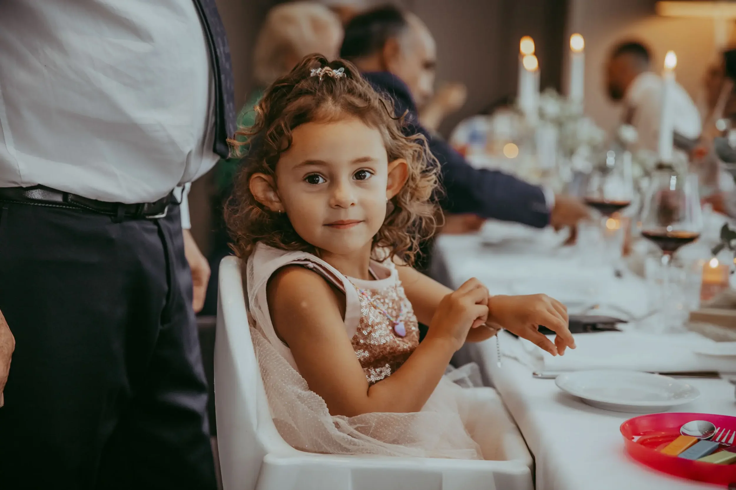 Ein junges Mädchen mit lockigem Haar sitzt an einem festlich gedeckten Tisch bei einem Familienessen, im Hintergrund sind weitere Menschen bei der Mahlzeit zu sehen.