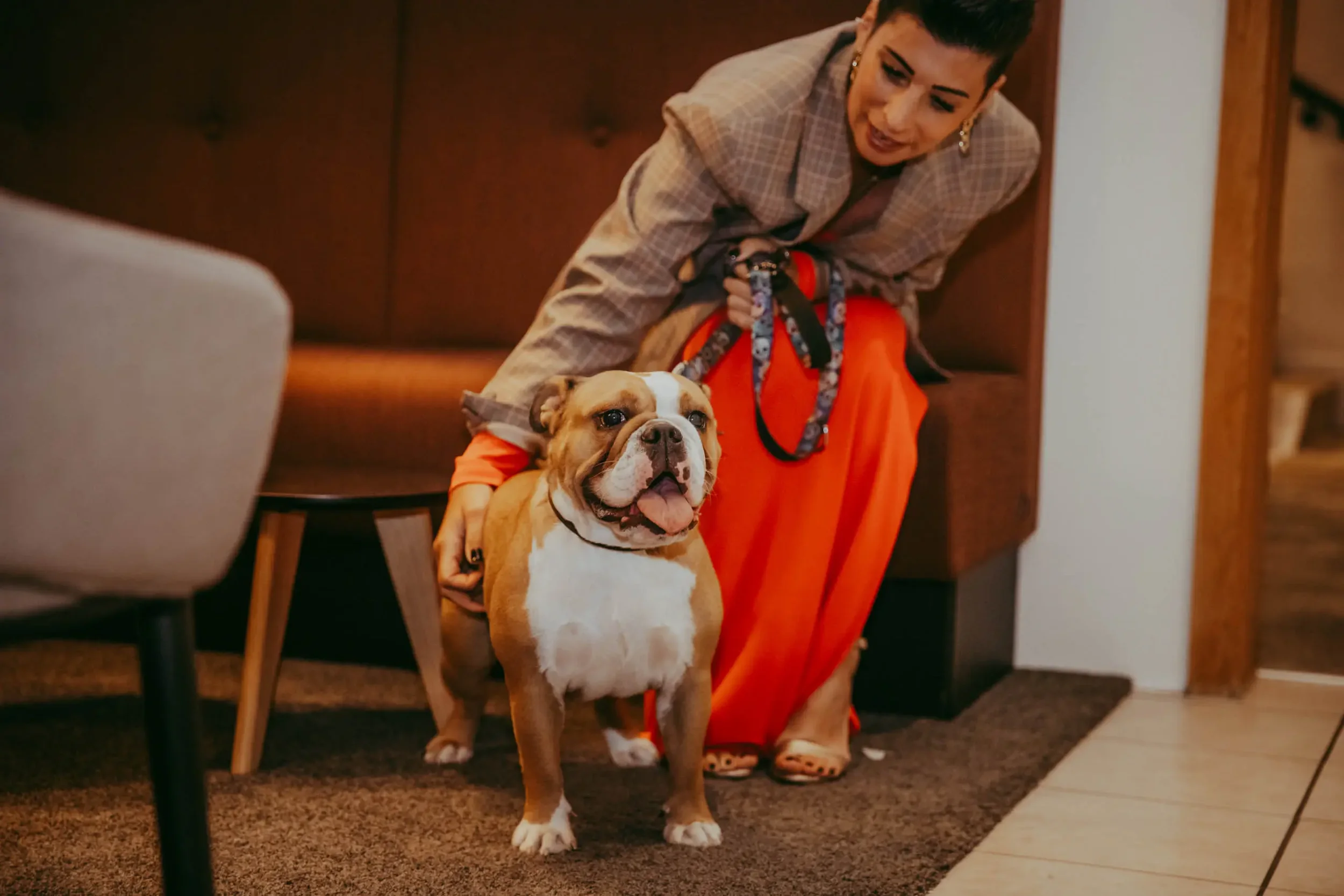 Eine Frau mit kurzen schwarzen Haaren, die einen Bulldogge-Hund festhält, der auf dem Boden steht. Die Frau trägt eine karierte Jacke und orange Hosen, während der Hund eine Leine trägt und in die Kamera blickt.