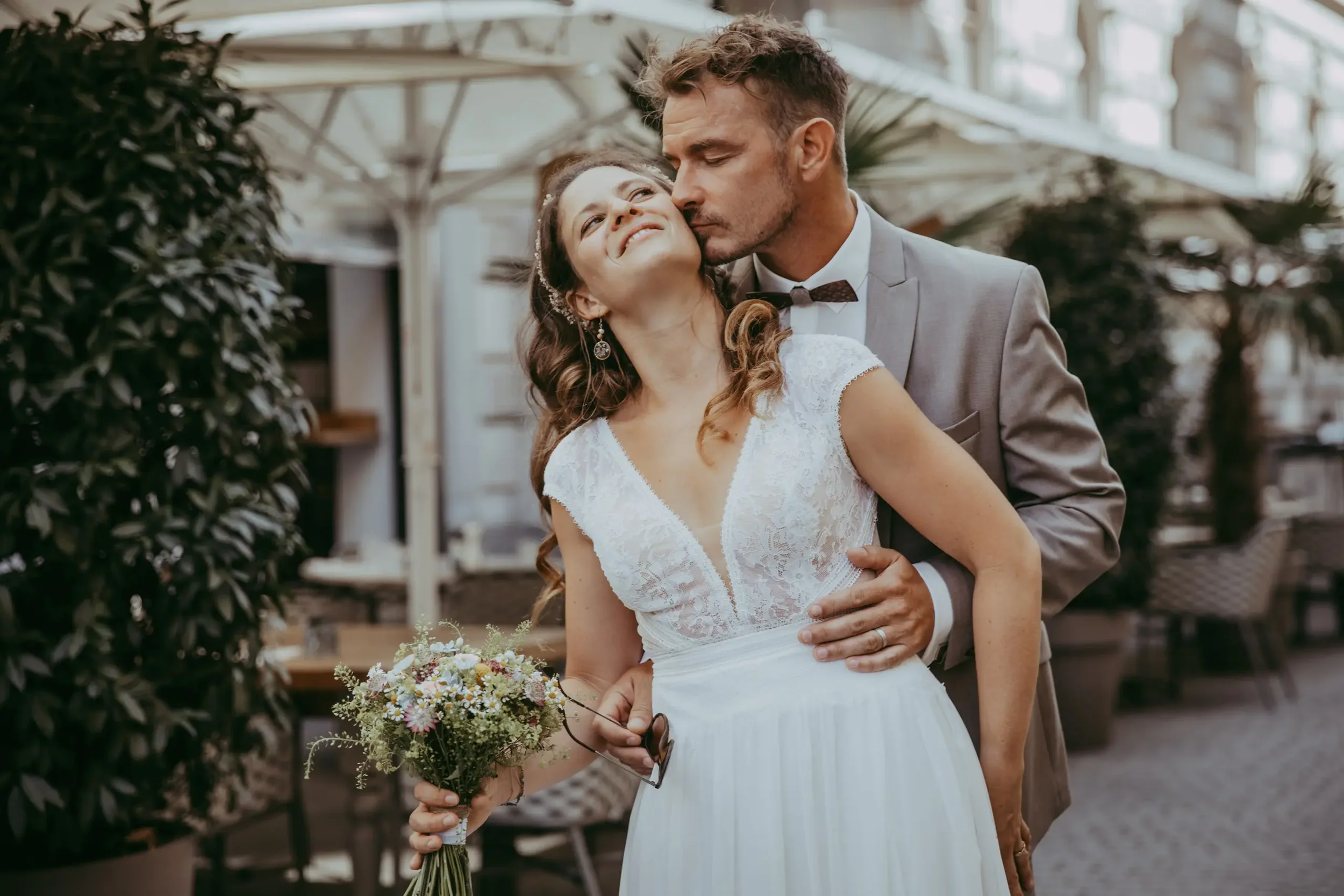 Ein Brautpaar bei einer Hochzeit, die Frau hält einen Blumenstrauß, der Mann küsst sie zärtlich im Freien.