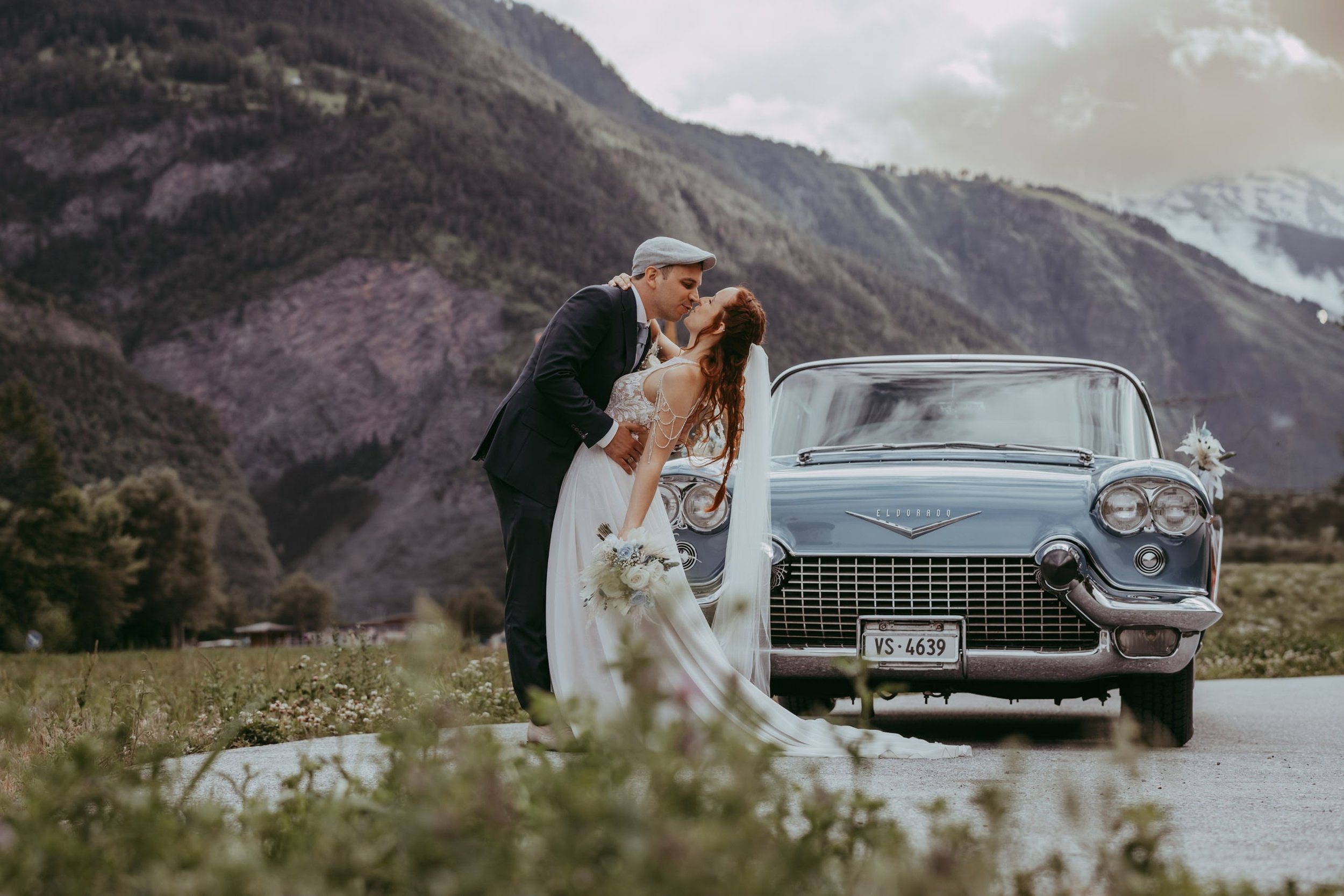 Ein Brautpaar küsst sich vor einem Vintage-Auto mit Berglandschaft im Hintergrund.