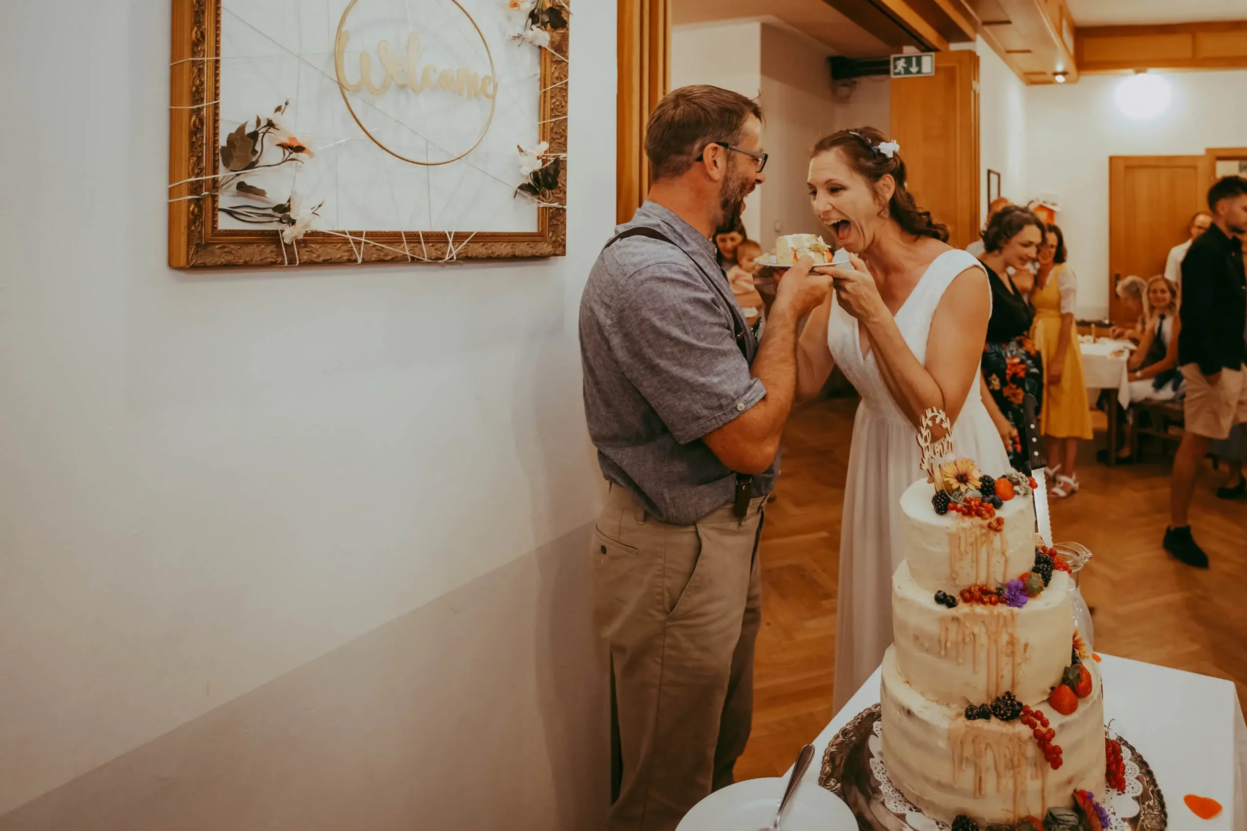 Ein Brautpaar scherzt mit einem Stück Hochzeitstorte bei einer Feier, im Hintergrund sind Gäste und ein Willkommen-Schild zu sehen.
