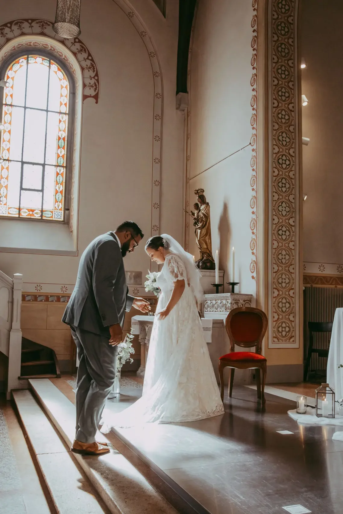 Braut und Bräutigam stehen in einer Kirche bei der Trauung, die Braut trägt ein langes weißes Kleid und hält einen Blumenstrauß, der Bräutigam trägt einen grauen Anzug.
