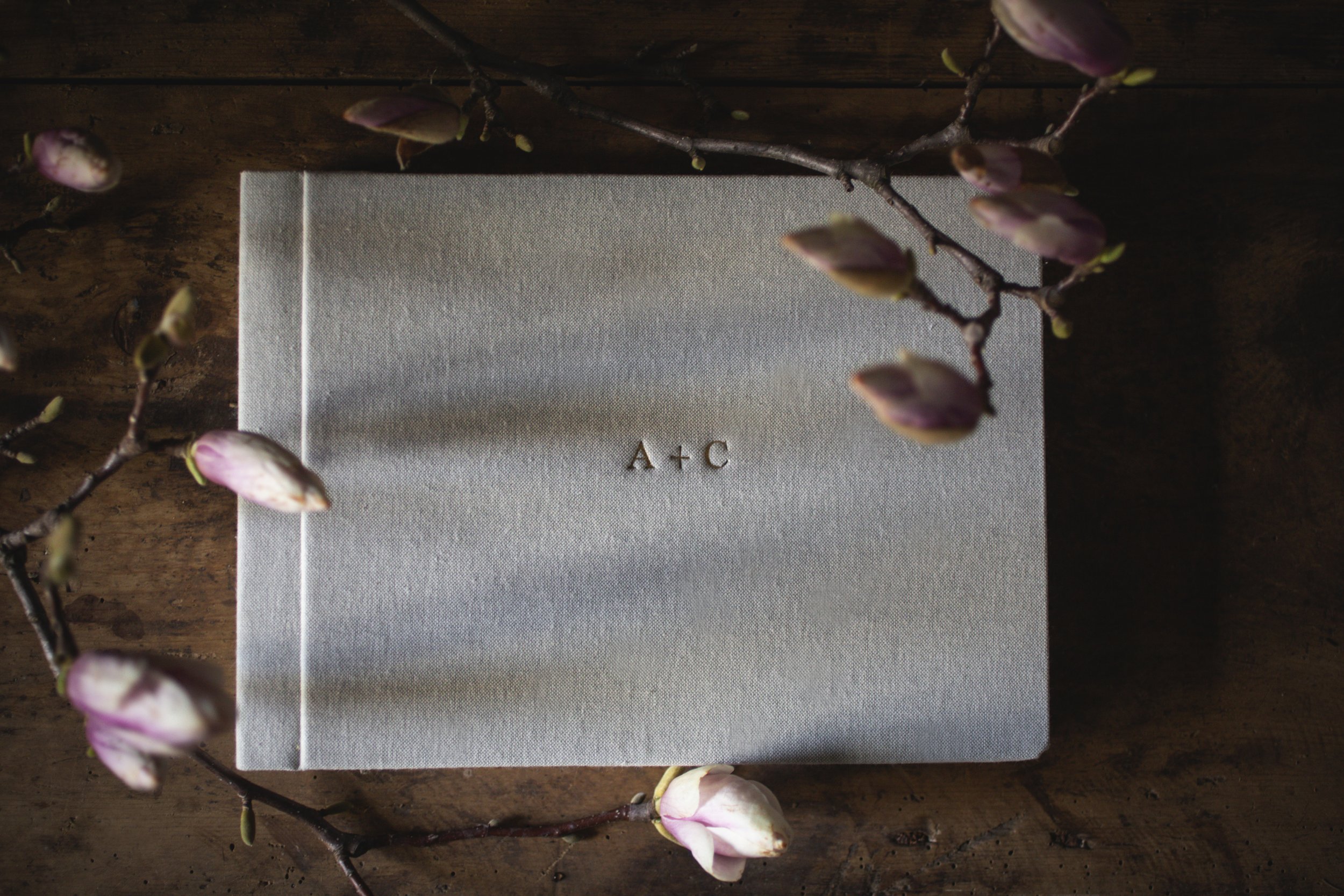 Ein weißes Buch mit den Buchstaben A + C auf dem Einband, umgeben von Magnolienzweigen mit pinken Knospen, liegt auf einem dunklen Holzuntergrund.