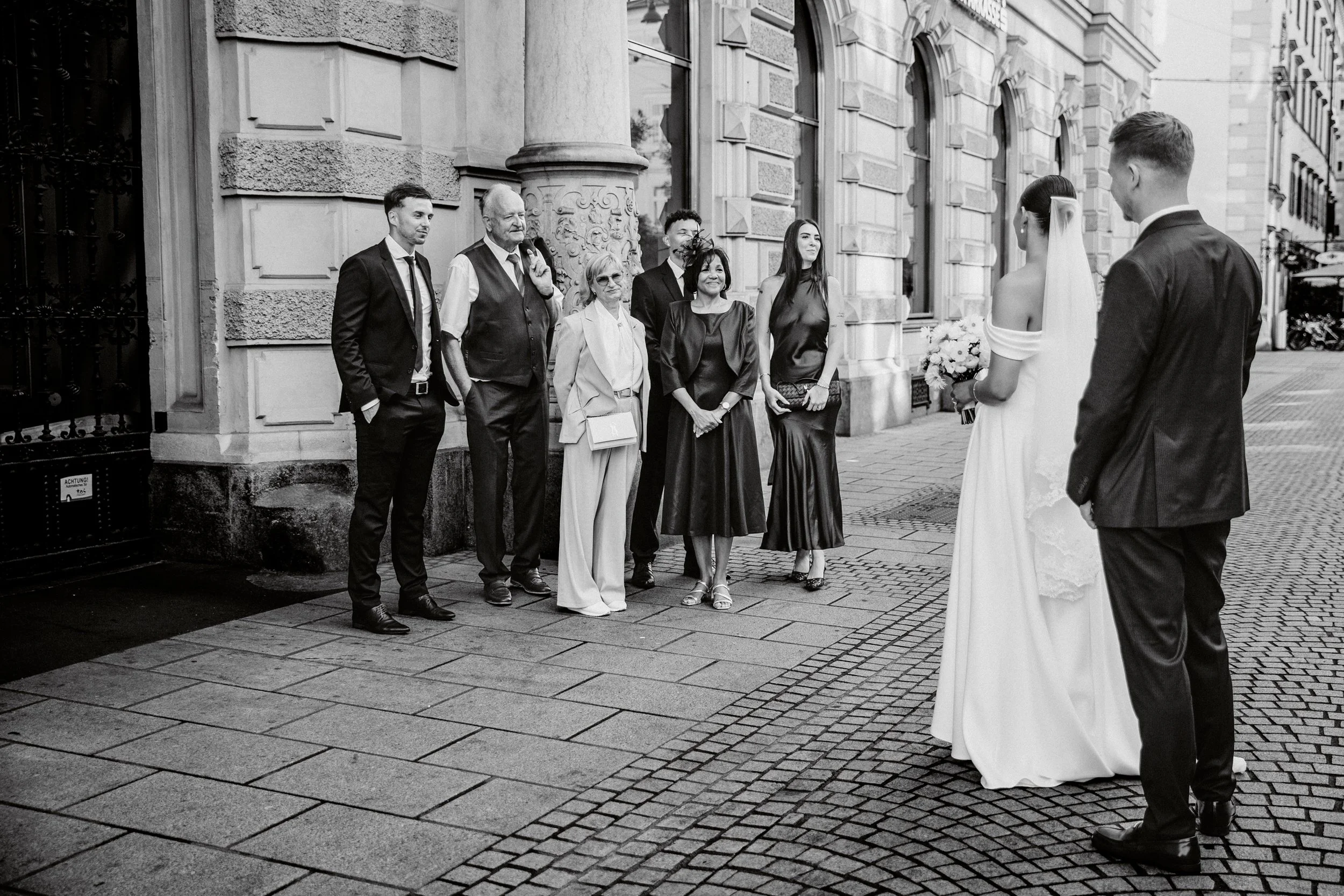 Eine Gruppe von Menschen, darunter Frauen und Männer, steht vor einem historischen Gebäude, während ein Brautpaar auf der rechten Seite vor ihnen steht. Es ist eine schwarze und weiße Fotografie, die möglicherweise eine Hochzeit oder ein besonderes E
