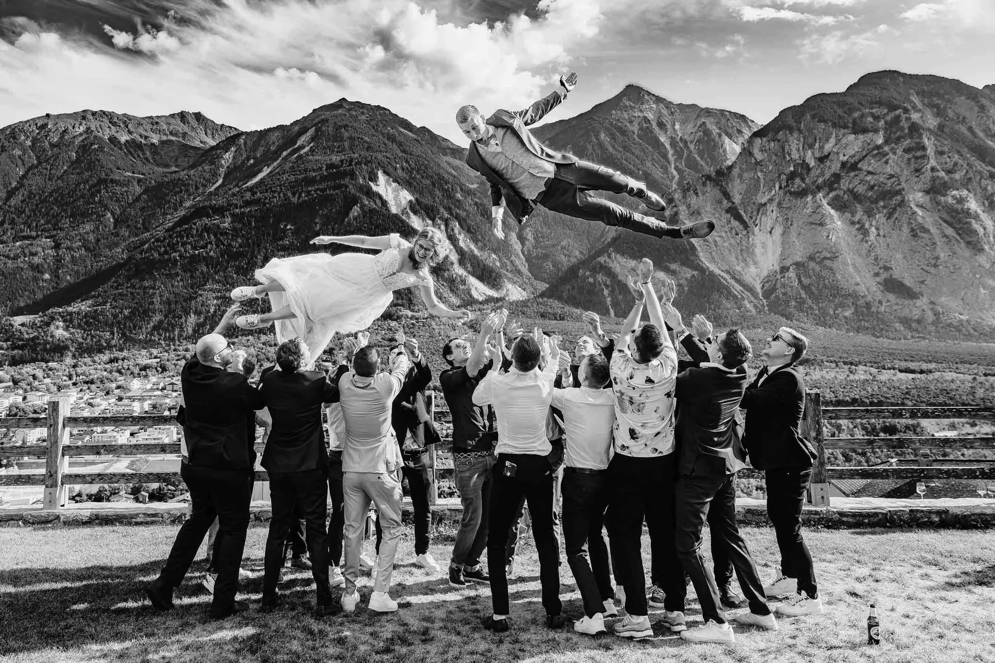 Eine Gruppe von Menschen wirft einen Mann in einem formellen Anzug in die Luft, während im Hintergrund Berge und Wolken zu sehen sind.