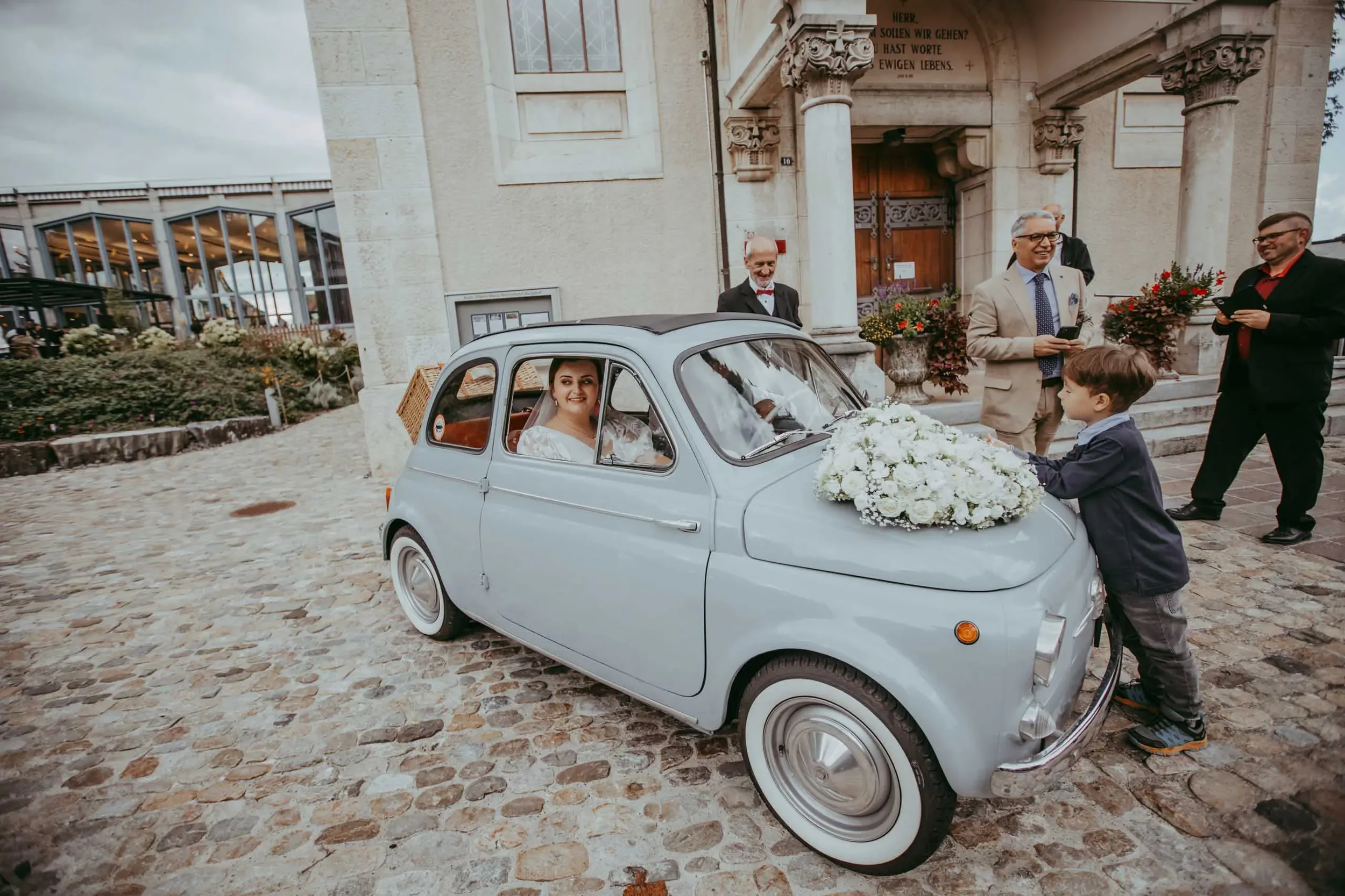 Braut in einem alten weißen Kleinwagen mit Blumen vor einer Steinarchitektur, umgeben von Männern in Anzügen, einer davon wird ein Kind in den Wagen schieben, während andere lachen.