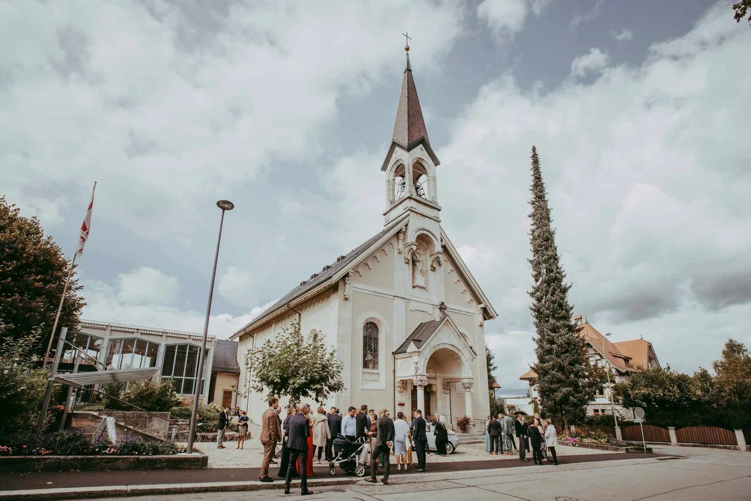 Eine weiße Kirche mit hoher steiler Dachspitze und Kirchenglocken, im Vordergrund versammelte Menschen bei einer Veranstaltung, umgeben von Bäumen und Gebäuden, bei bewölktem Himmel.