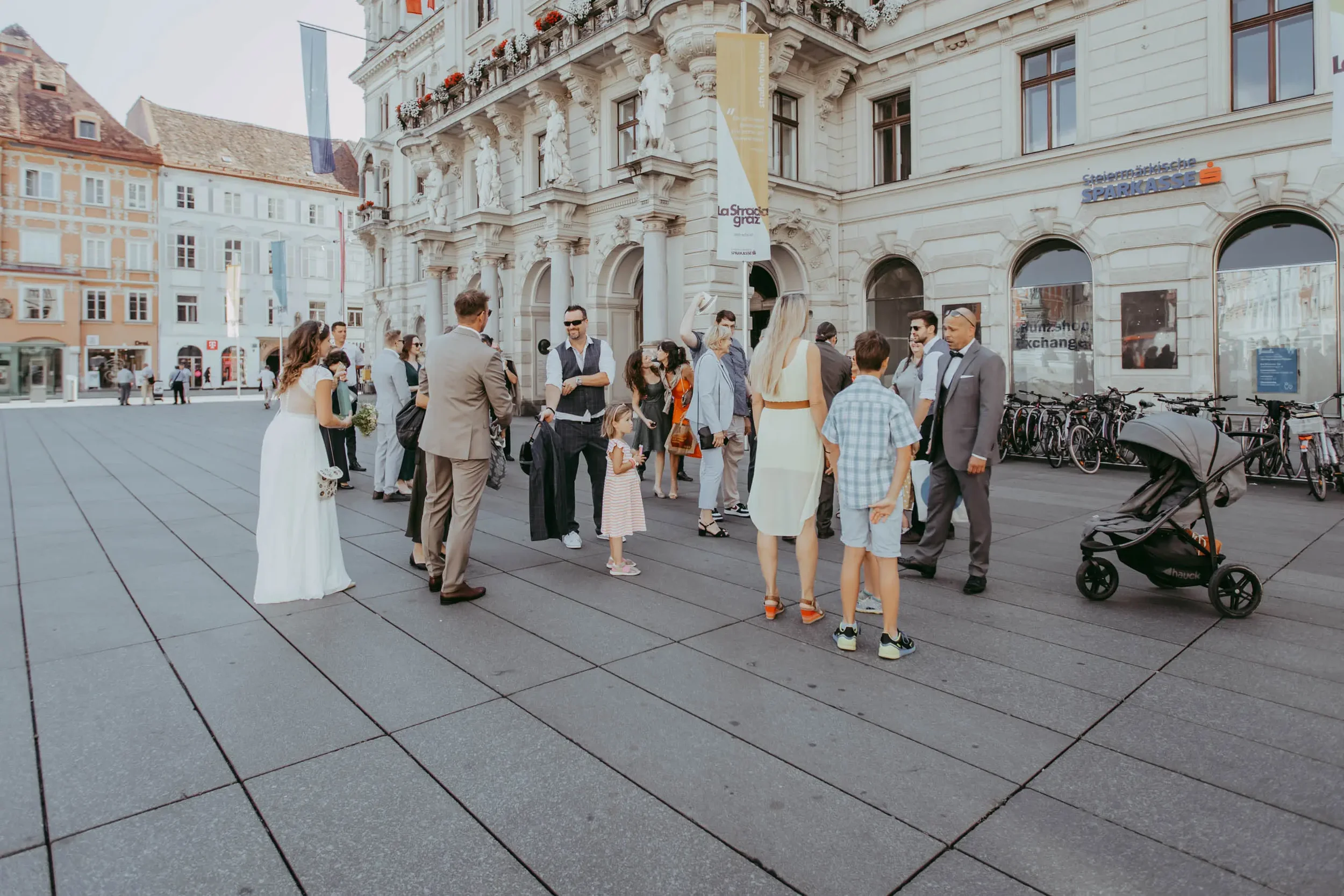 Menschen versammeln sich vor einem historischen Gebäude in einer Stadt, einige tragen formelle Kleidung, darunter ein Mann im Anzug, eine Frau in einem weißen Kleid, und ein Kind mit rotem gestreiftem Kleid. Mehrere Fahrräder sind an der Gebäudewand 