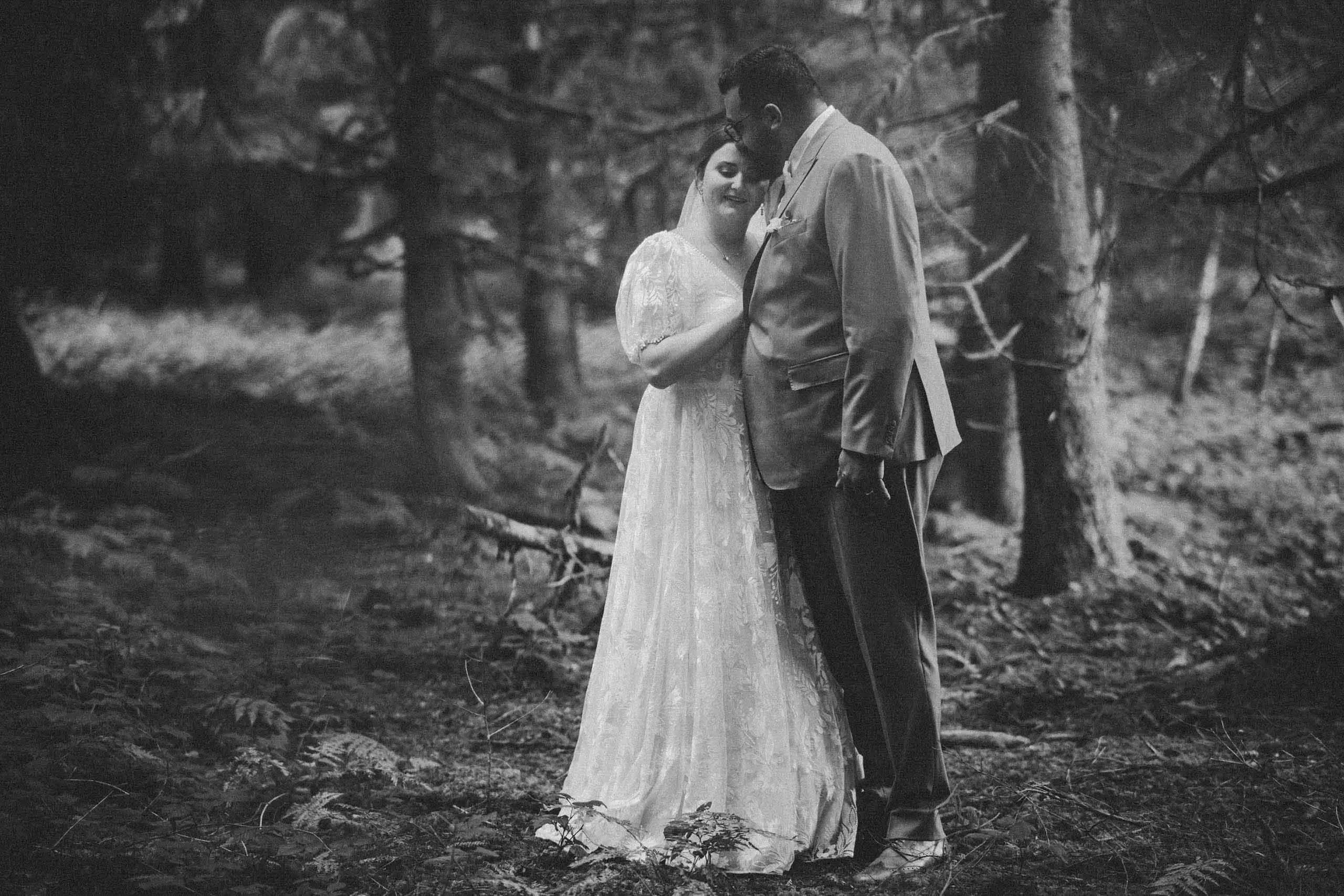 Schwarz-weiß-Foto eines Paares an ihrer Hochzeit in einem Wald, umarmend und lächelnd, mit Bäumen im Hintergrund.