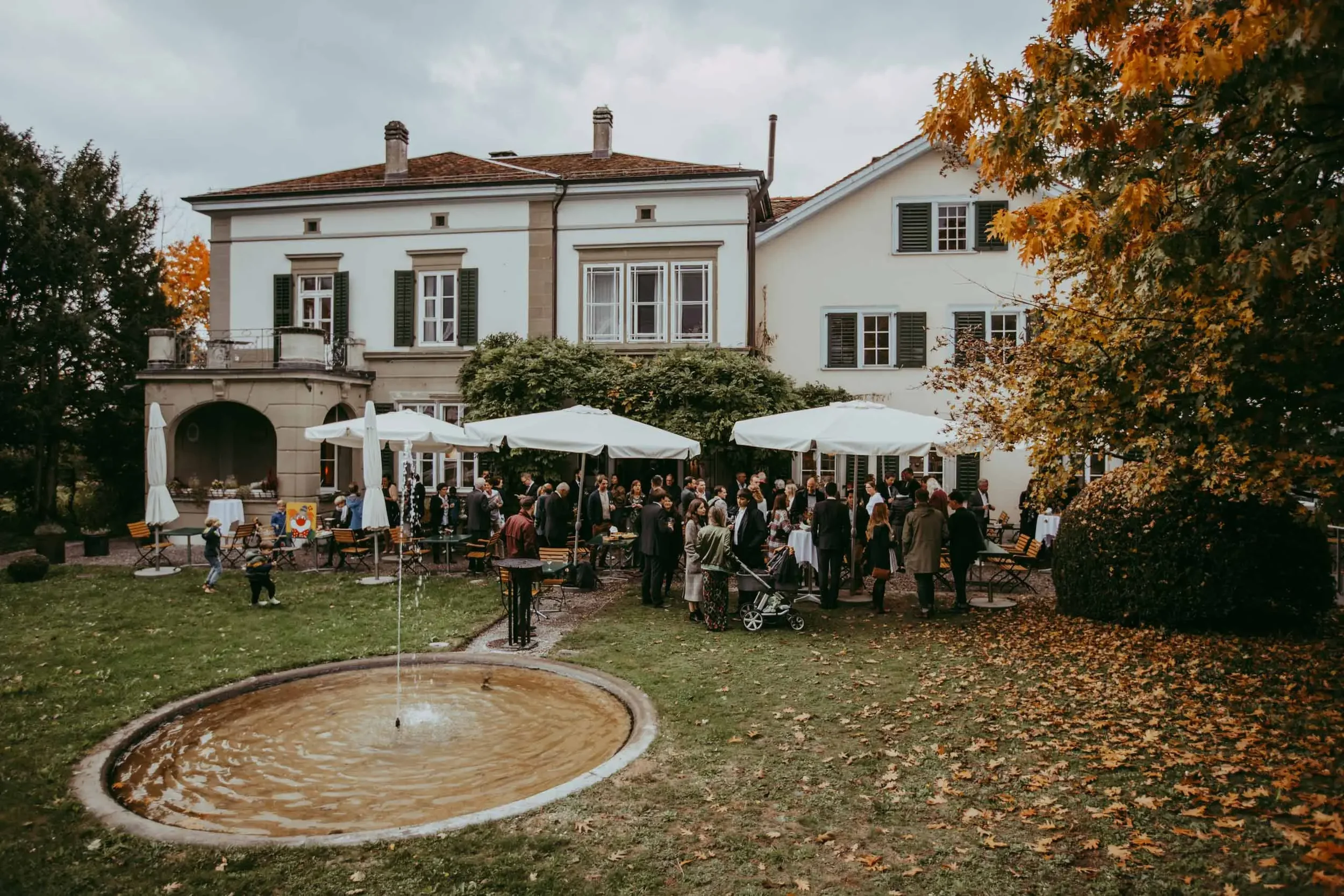 Bauernhaus mit Terrasse, umgeben von Bäumen mit Herbstlaub, voller Menschen bei einem Fest oder Empfang, im Vordergrund ein Brunnen mit Wasser, im Hintergrund grauen Wolken am Himmel.