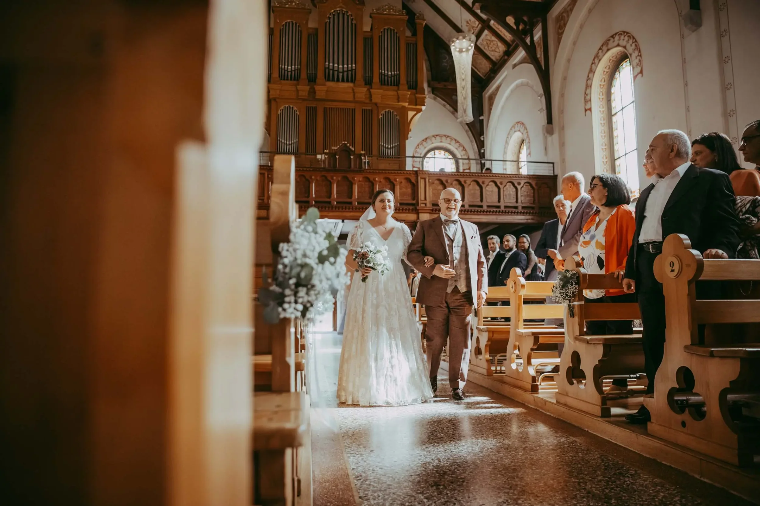 Braut und Vater bei einer Hochzeit in einer Kirche, umgeben von Gästen, Blick von einer Nische durch ein Holztor