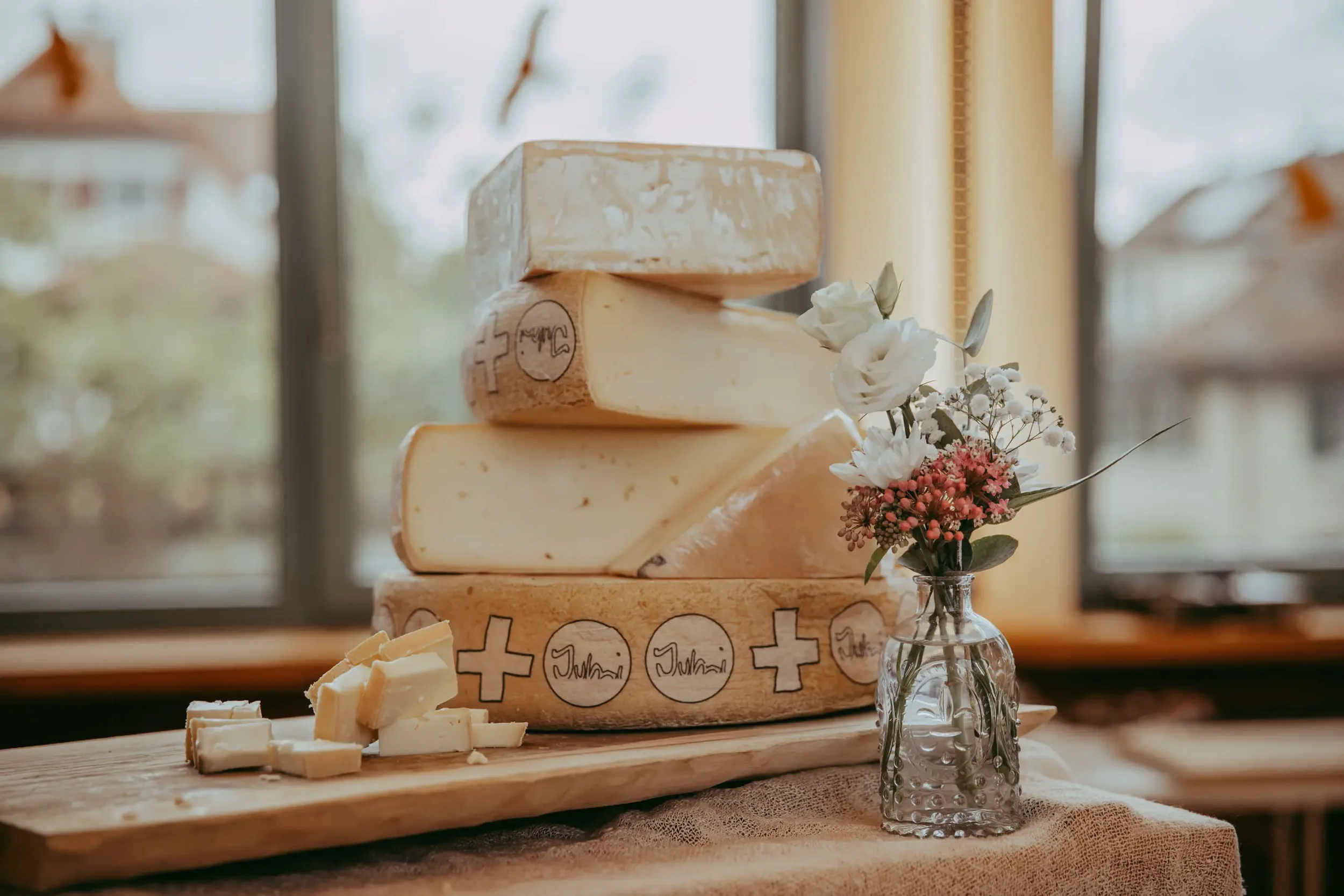 Käse auf einem Holzbrett, eingeritzt mit Kreuzen und Namen, daneben eine kleine Vase mit weißen und rosa Blumen, vor einer Fensterkulisse.