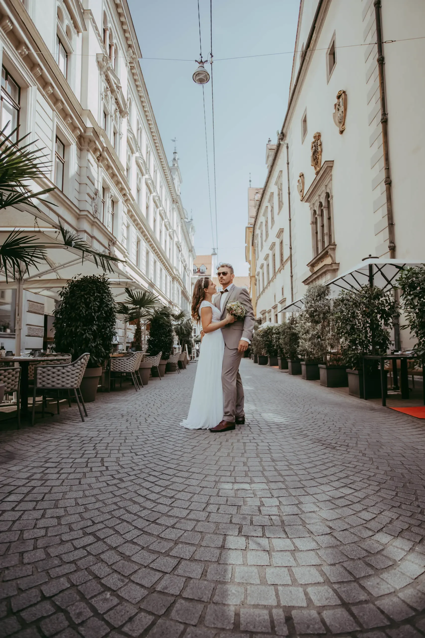 Ein Brautpaar bei einer Hochzeit auf einer gepflasterten Straße, umgeben von historischen Gebäuden und Outdoor-Gastronomie. Die Frau trägt ein weißes Brautkleid und hält einen Brautstrauß, der Mann trägt einen grauen Anzug mit Fliege.