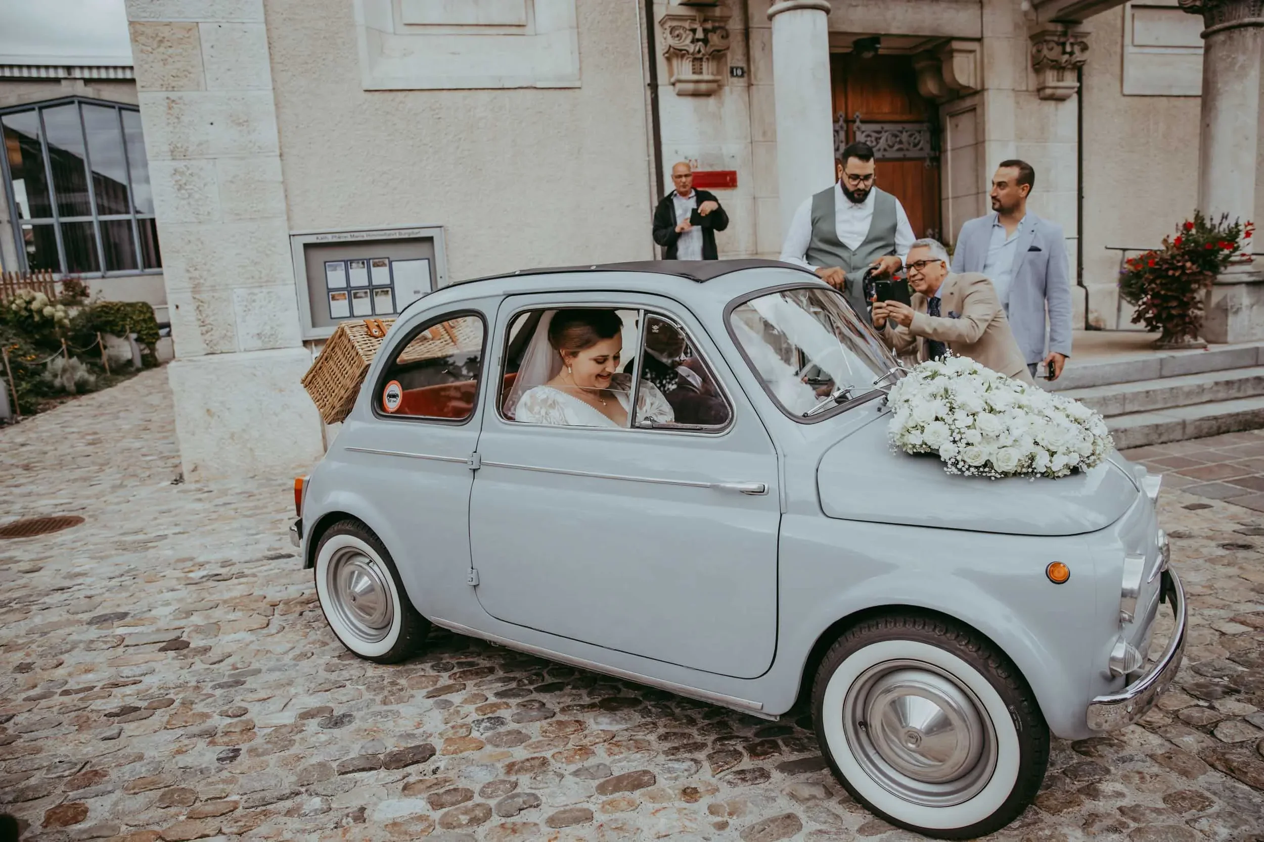 Ein weißer Oldtimer mit einem Blumenarrangement auf der Motorhaube und einem Brautpaar im Auto vor einer alten Steinfassade, umgeben von mehreren Personen, die Fotos machen.