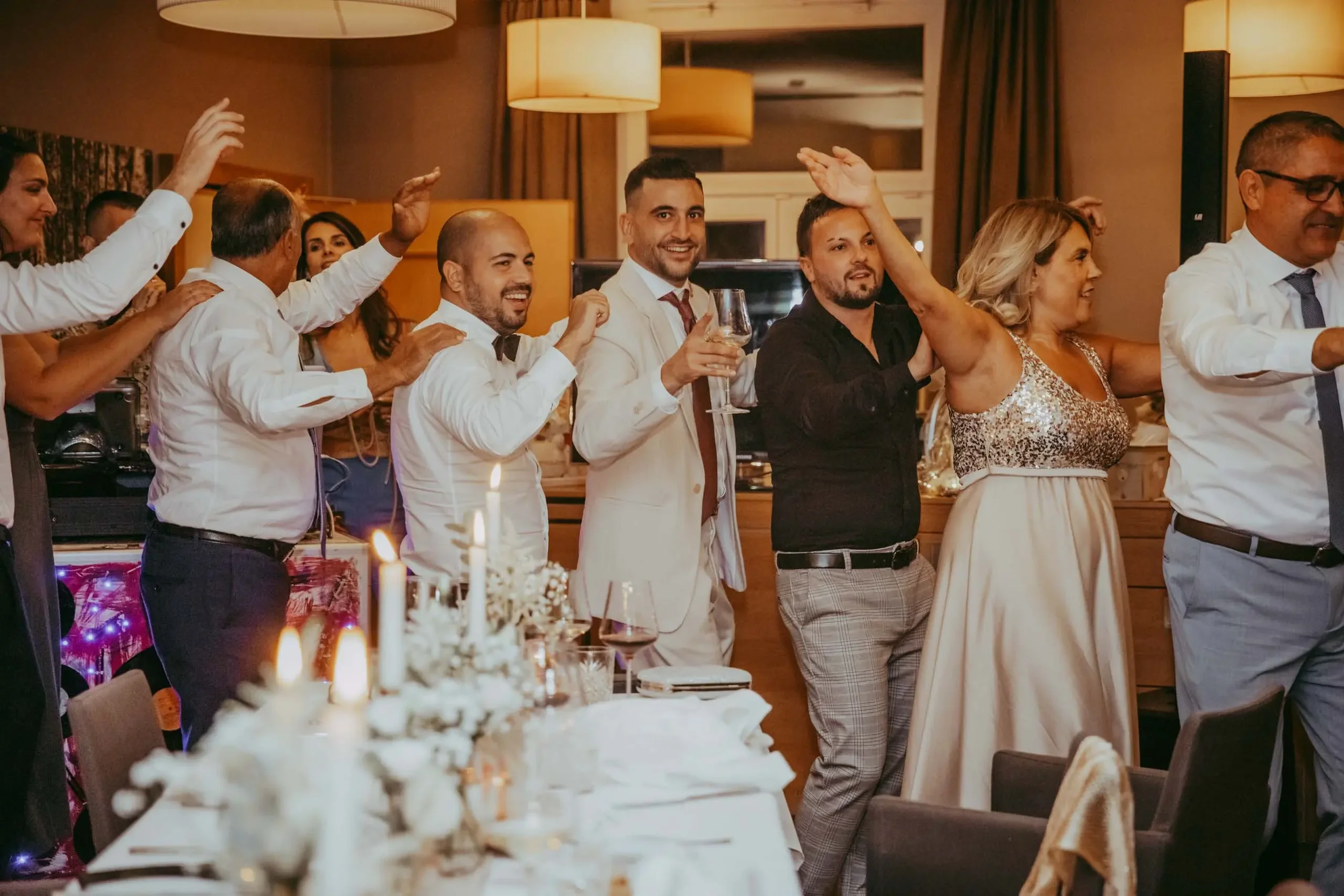 Menschen feiern bei einer Party oder einem Empfang, einige tanzen und einige halten Gläser in der Hand, in einem festlich dekorierten Raum mit Kerzen auf einem Tisch im Vordergrund.