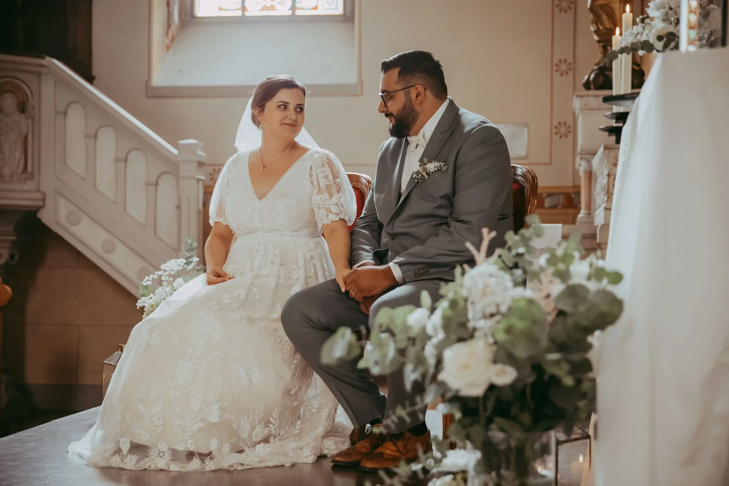Ein Brautpaar sitzt in einer Kirche während einer Hochzeitszeremonie, die Braut trägt ein weißes Hochzeitskleid und der Bräutigam einen grauen Anzug.