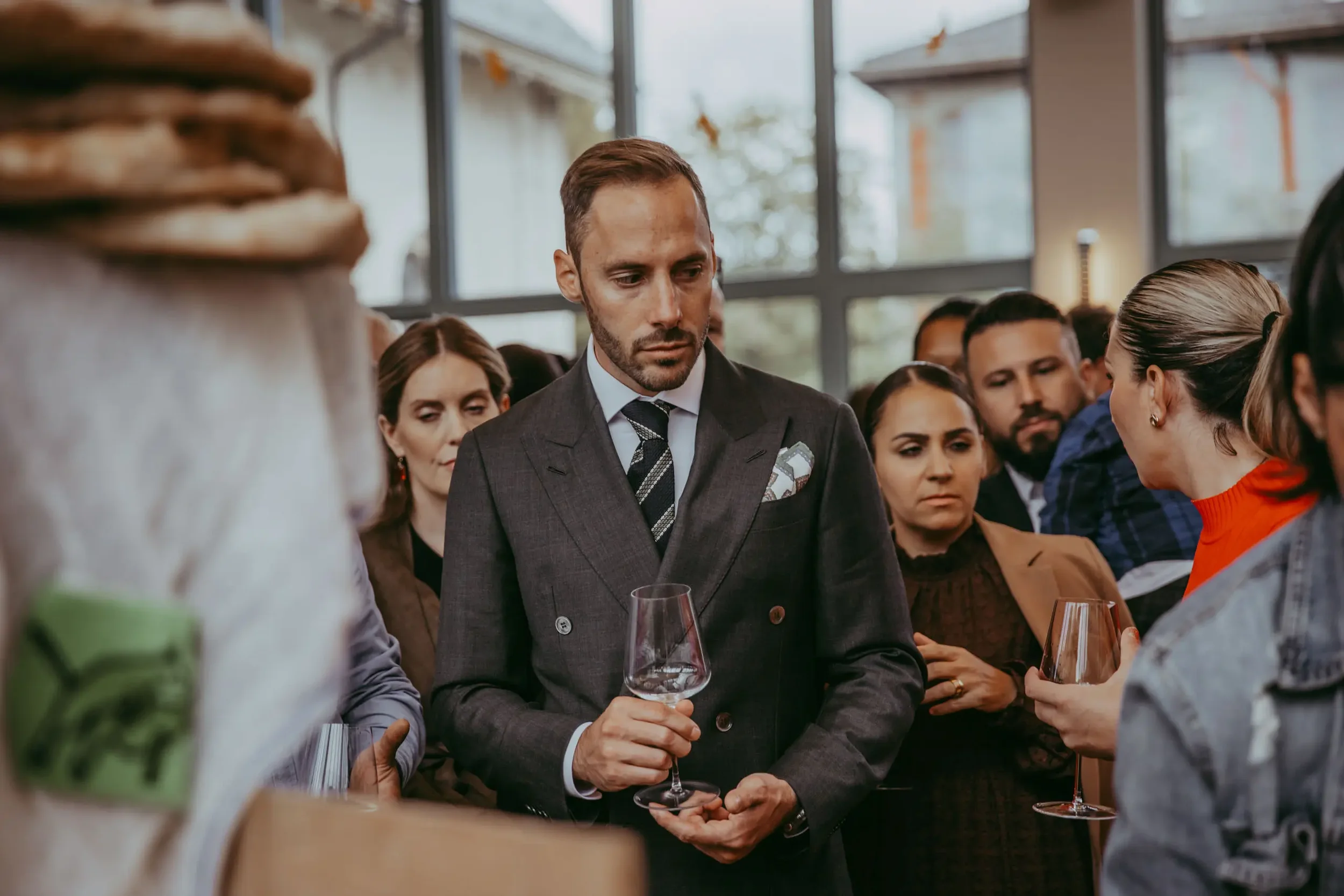 Ein Mann in einem grauen Anzug hält ein Glas Rotwein und unterhält sich bei einer Veranstaltung mit mehreren anderen Menschen.