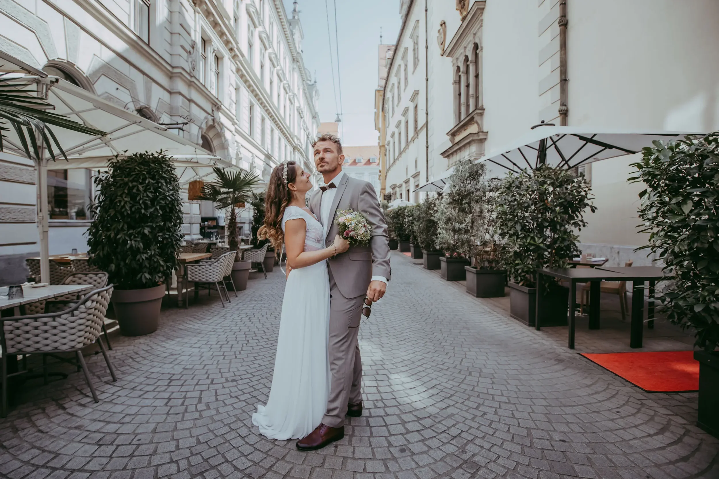 Ein Brautpaar in formeller Kleidung steht eng beieinander auf einer gepflasterten Straße in einer Altstadt mit historischen Gebäuden, umgeben von Cafés und Pflanzen, bei Tageslicht.
