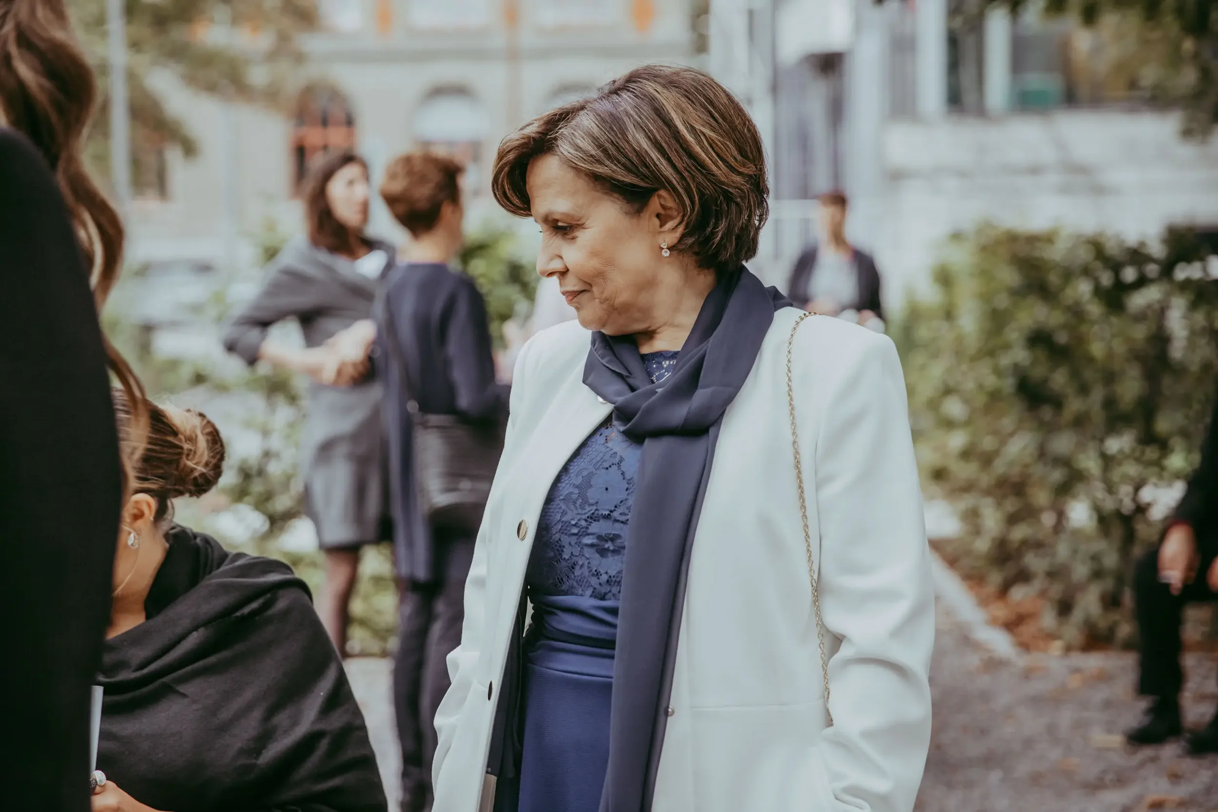 Eine ältere Frau in weißer Jacke und blauer Bluse, die mit einer jüngeren Person spricht, während andere Menschen im Hintergrund draußen unterwegs sind.