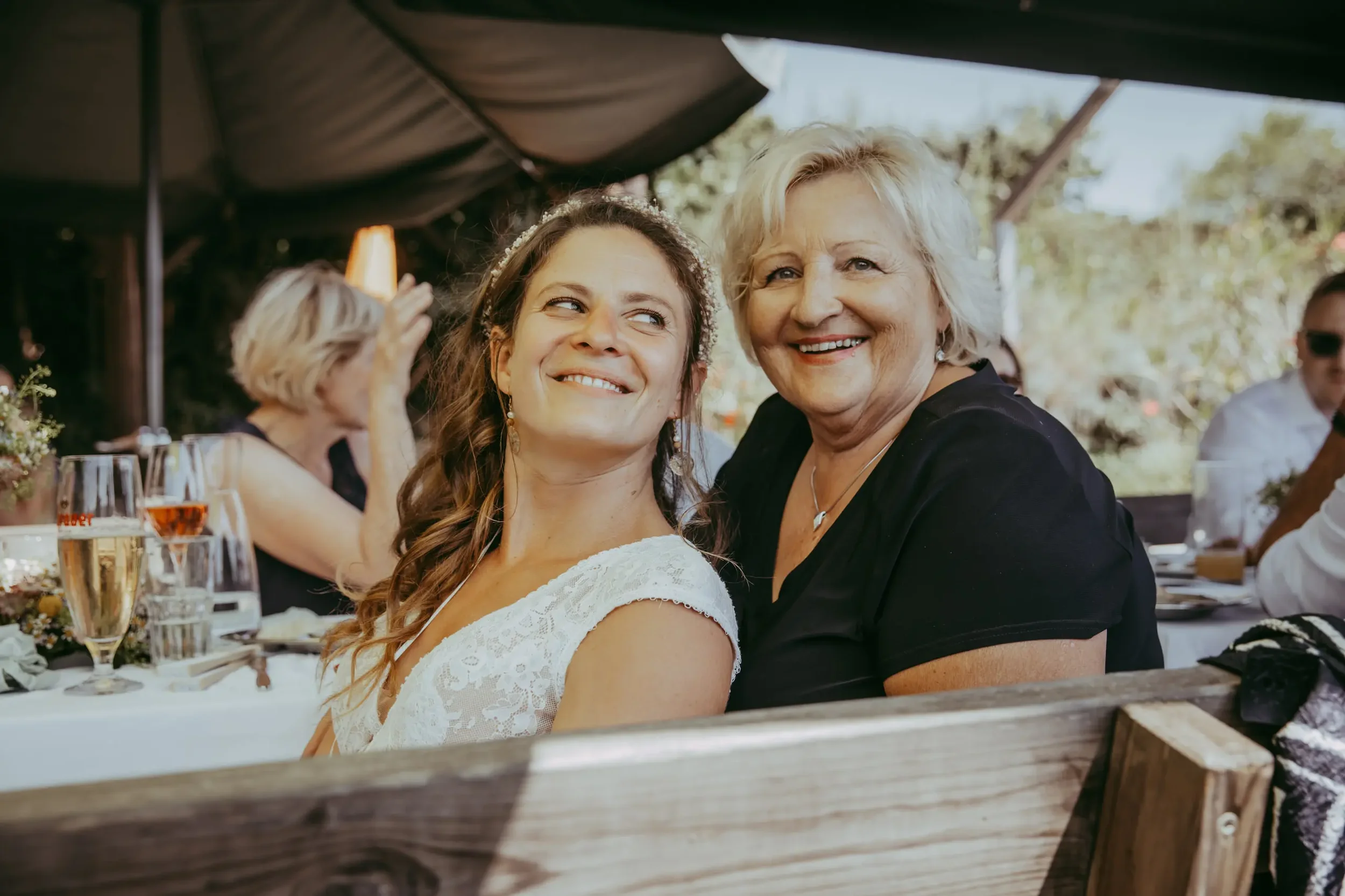 Eine junge Frau in einem weißen Kleid und eine ältere Frau in schwarzer Kleidung sitzen bei einer Feier im Freien unter einem großen Sonnenschirm, lachen und schauen sich liebevoll an.