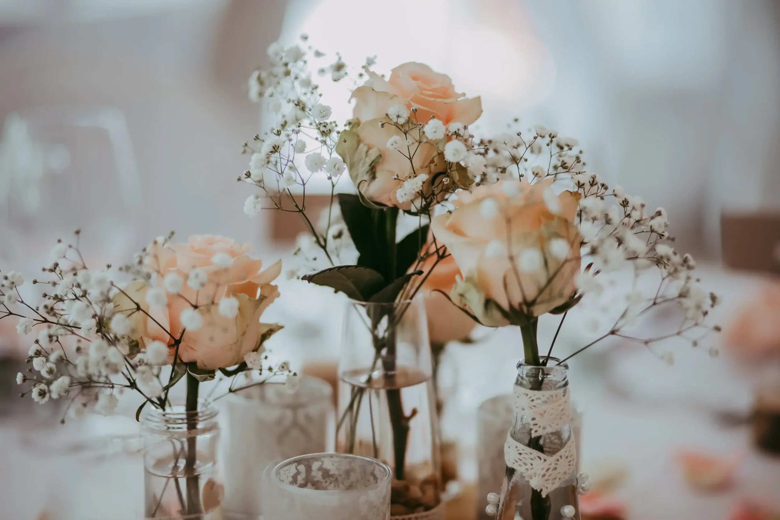 Blumen in Glasvasen, hauptsächlich mit Rosen und kleinen weißen Blumen, auf einem Tisch bei einer Feier.