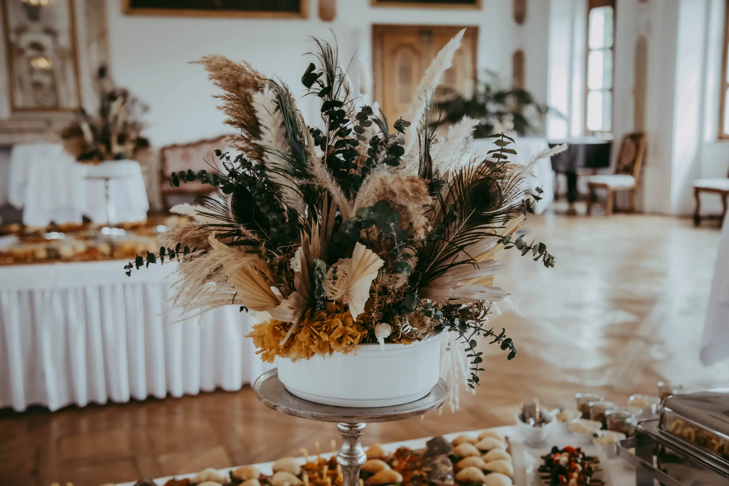Blumenstrauß mit Pampasgras und grünen Zweigen in einer weißen Vase auf einem hohen Glasständer, im Hintergrund ein Raum mit großen Fenstern und Vintage-Möbeln.