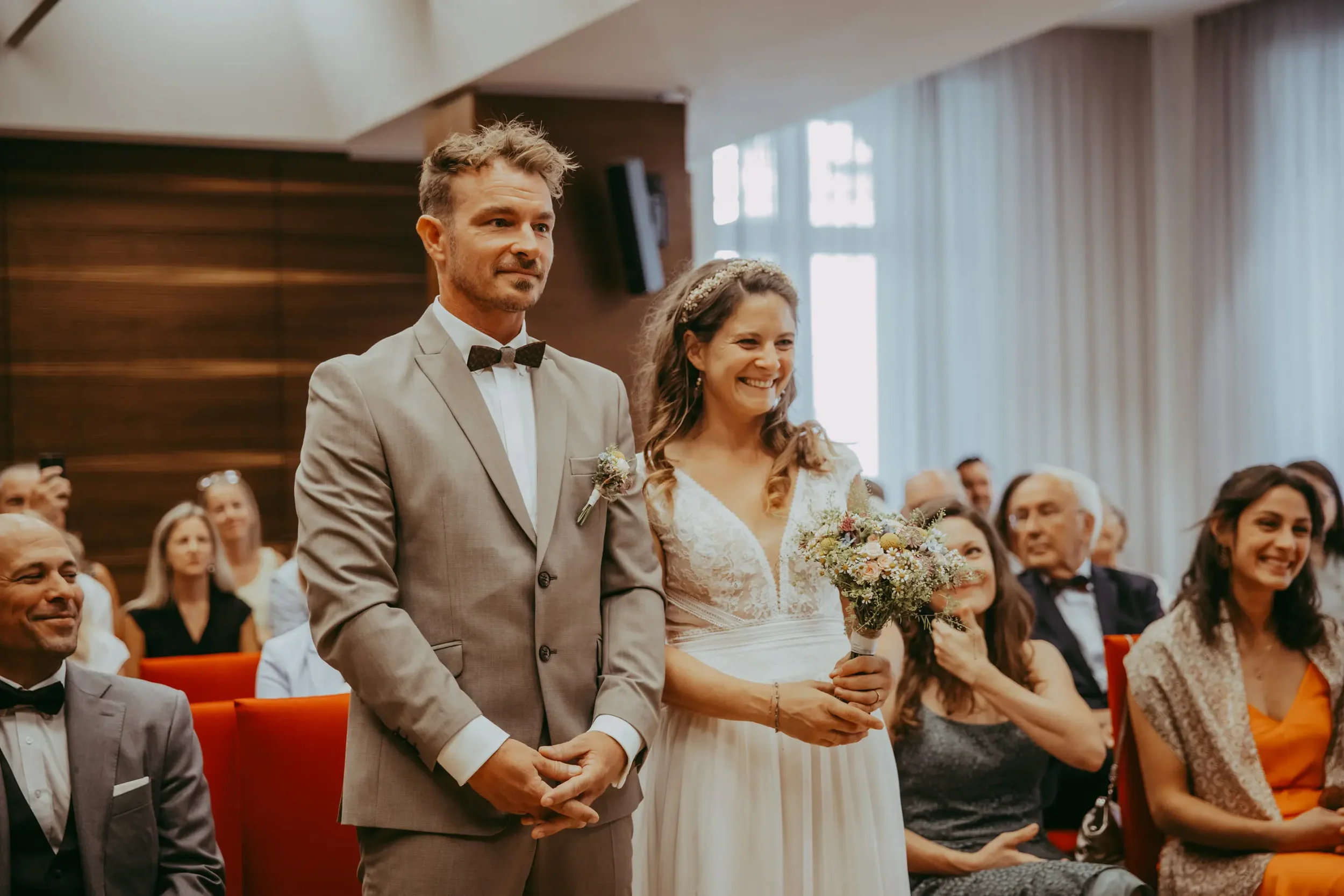 Ein Mann und eine Frau bei einer Hochzeitszeremonie stehen nebeneinander, während sie von einer Gruppe von Gästen beobachtet werden.