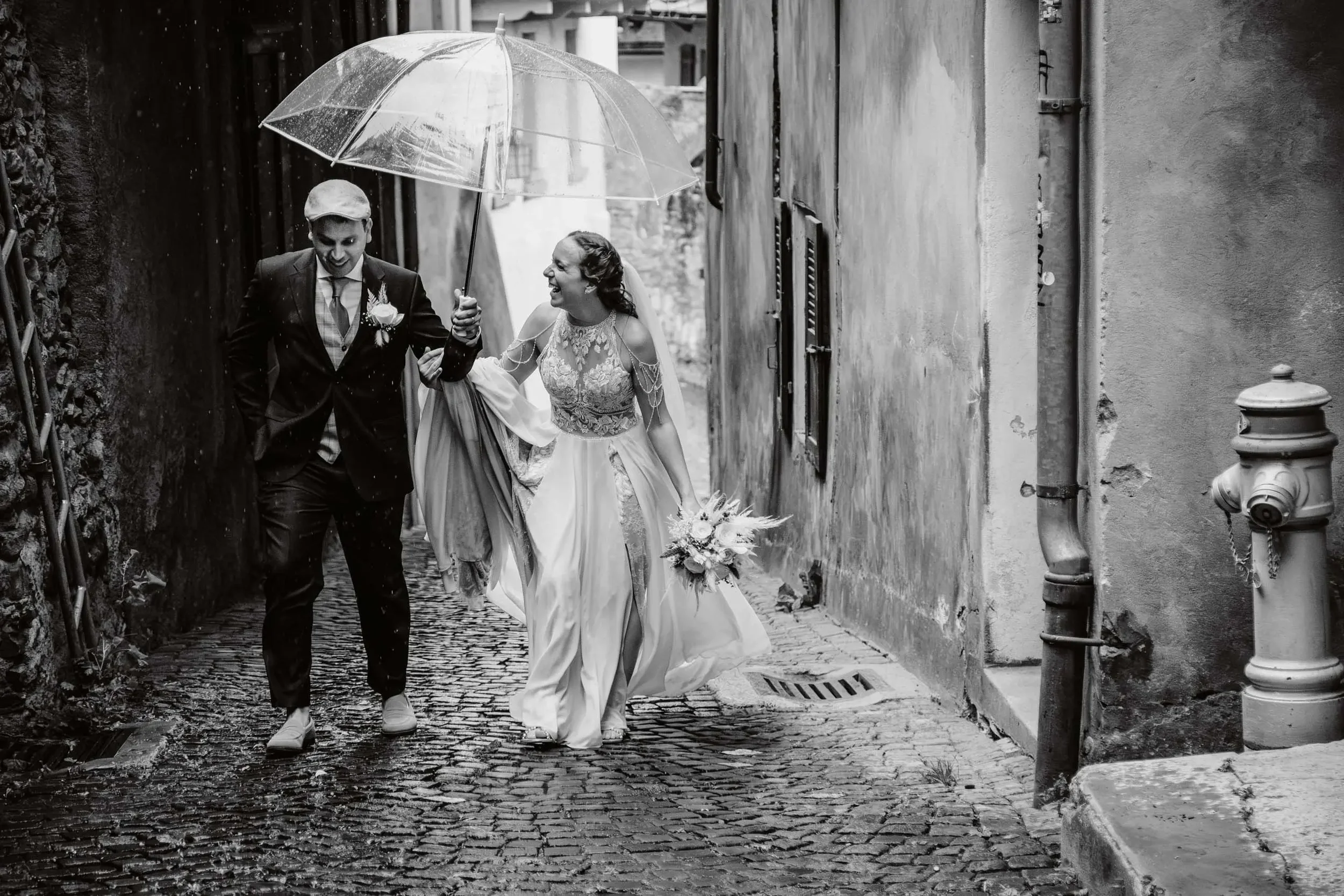 Ein lachendes Brautpaar spaziert arm in arm durch eine enge Gasse, während es einen Regenschirm hält, Wasser spritzt auf den Boden. Es regnet leicht, die Szene ist schwarz-weiß.