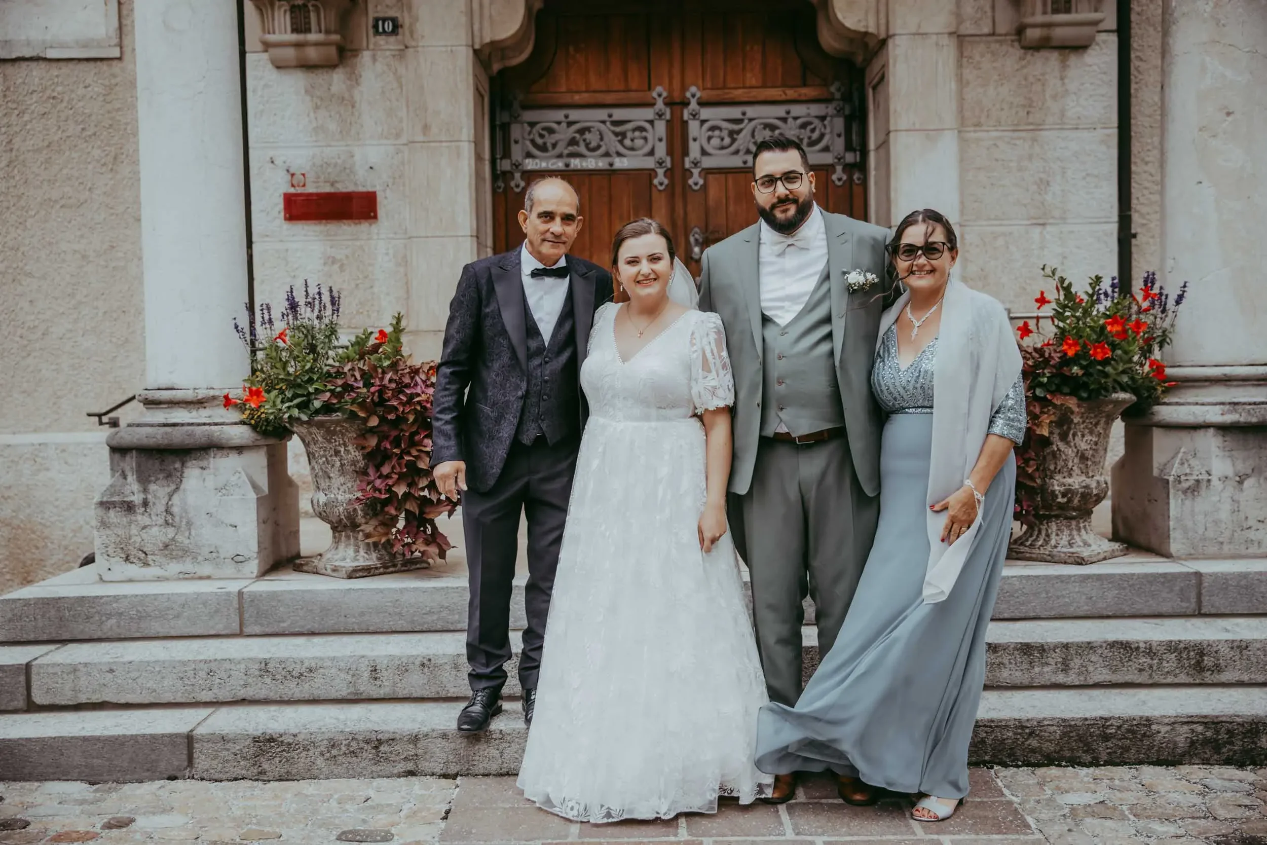 Eine Gruppe von fünf Menschen steht vor einer alten Stein- und Holztür, wobei zwei Männer und drei Frauen zu sehen sind. Es ist ein festlicher Anlass, wahrscheinlich eine Hochzeit, da eine Frau in einem weißen Kleid trägt und eine andere in einem ele